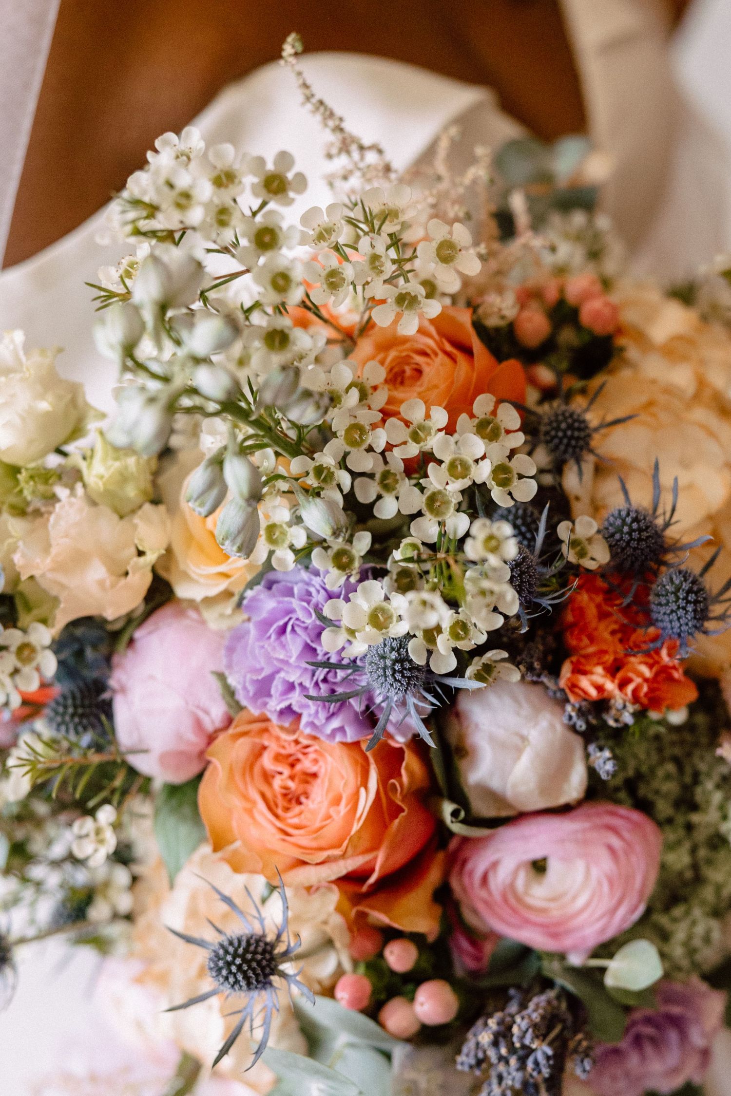 Lush bridal bouquet featuring orange roses, ranunculus, purple blooms, and white waxflowers.