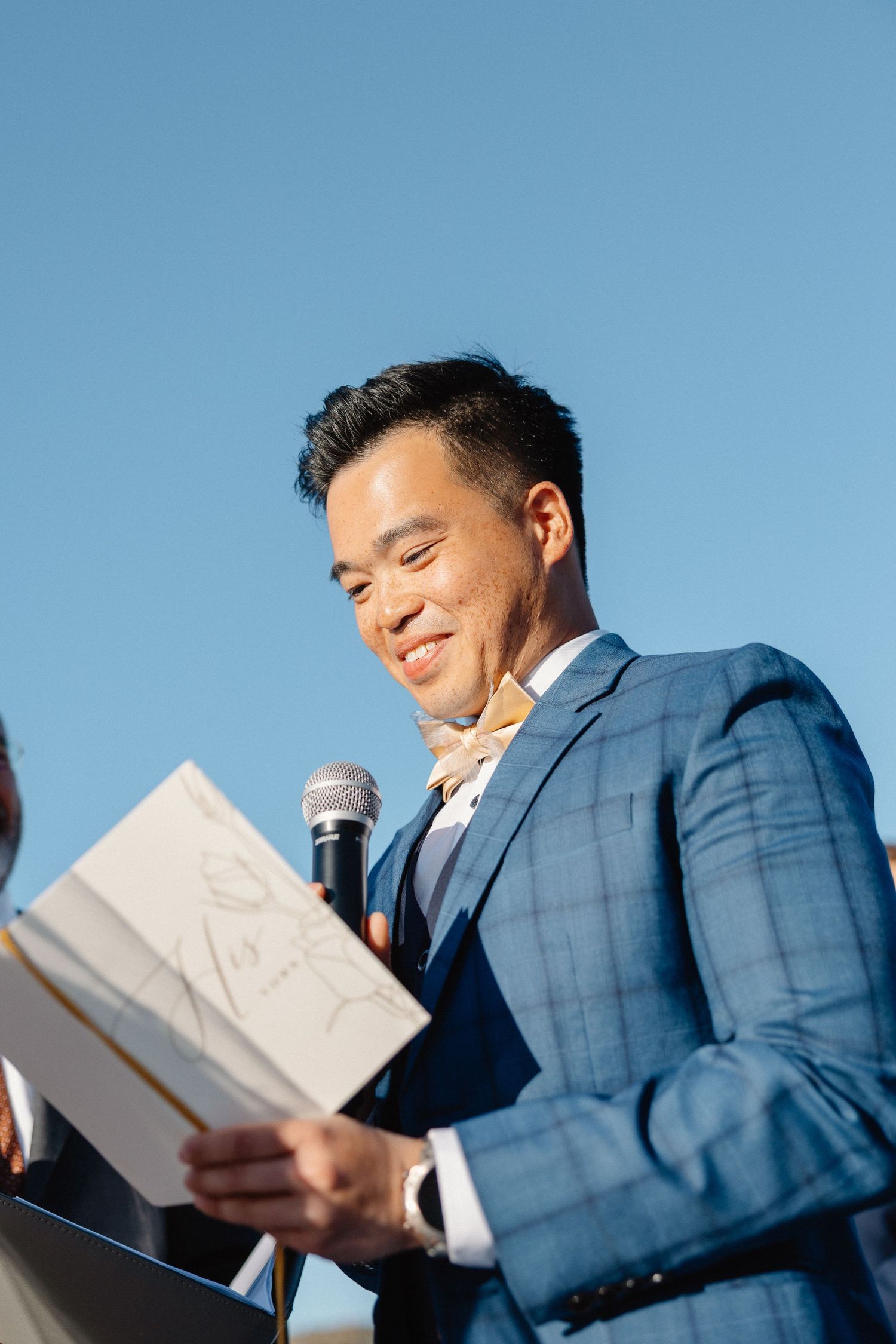 Man in blue suit smiling while reading vows from a card at an outdoor wedding ceremony.