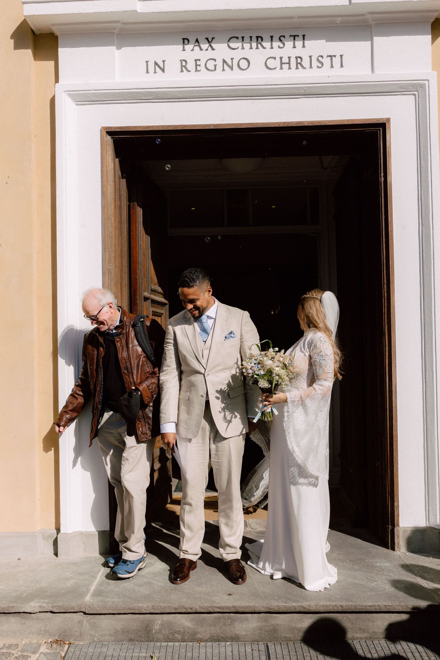 Wedding party poses outside church entrance with PAX CHRISTI inscription above.