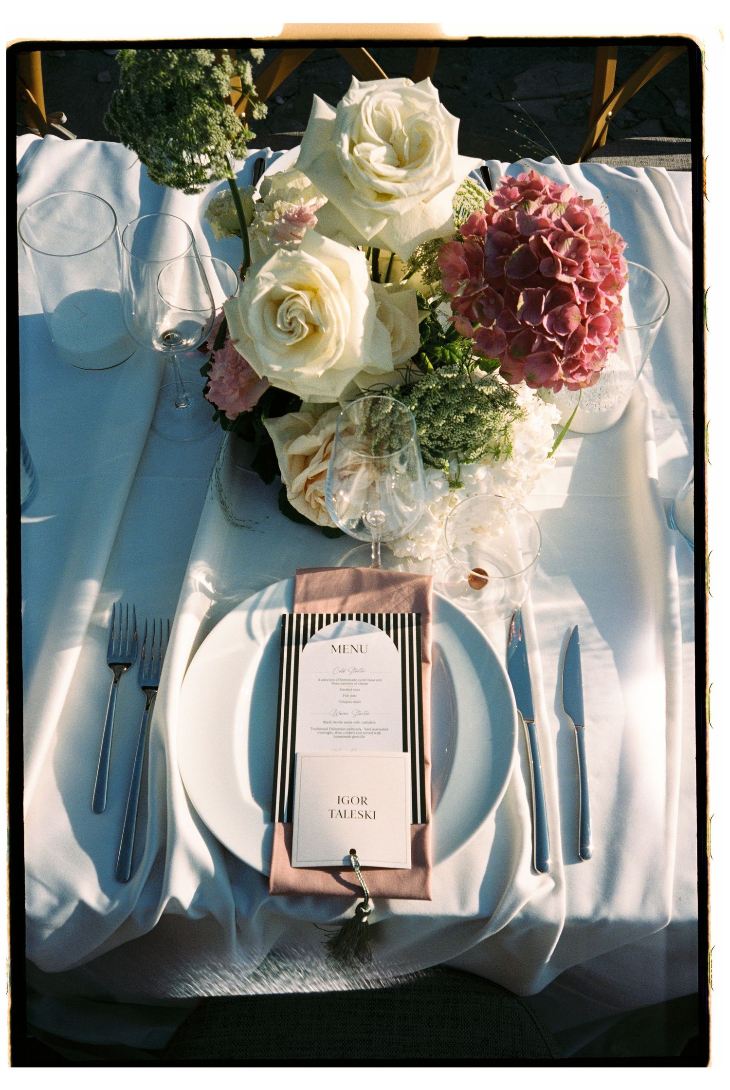 Elegant wedding table setting with white roses, pink hydrangeas, striped menu card, and polished silverware on white linen.