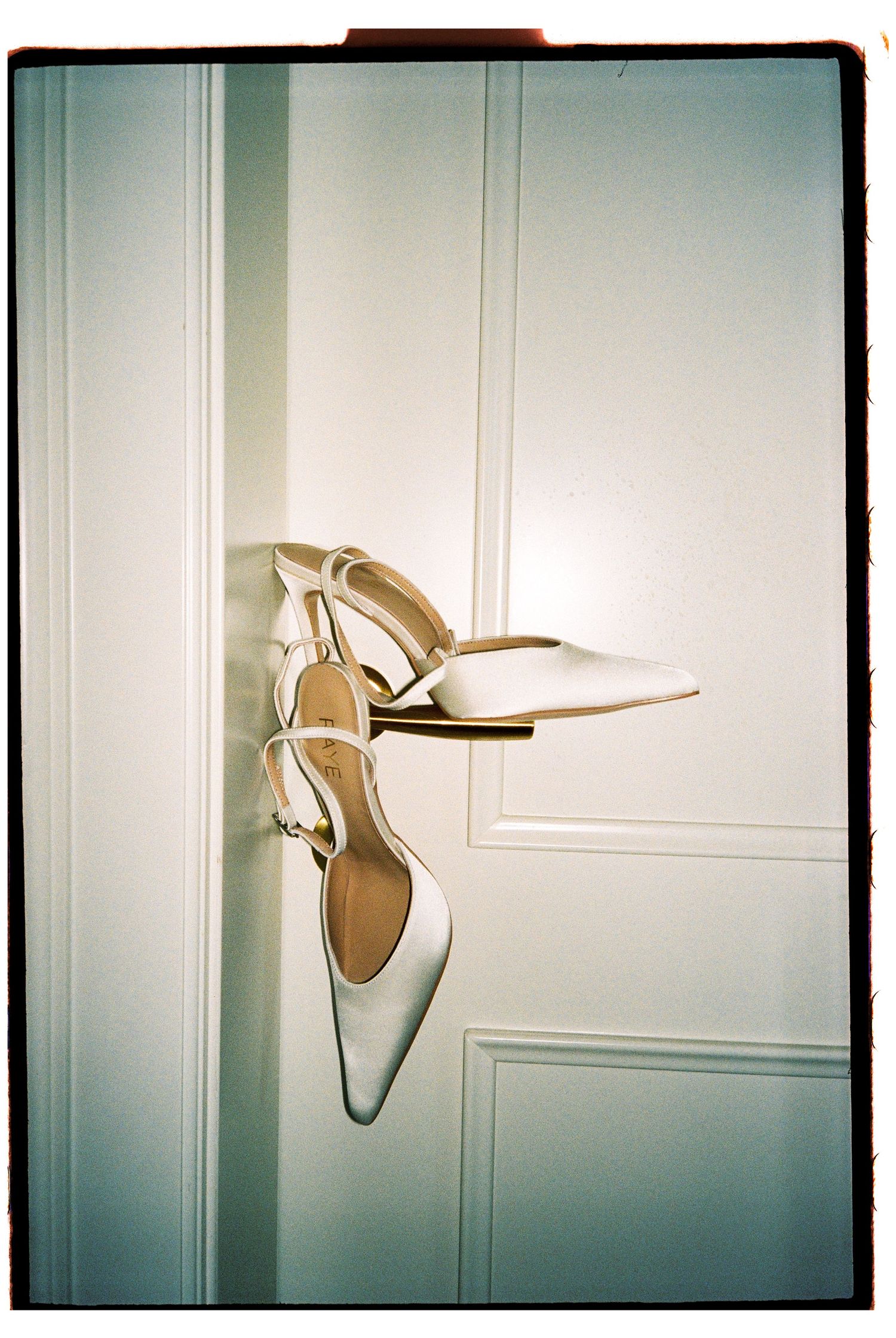 White slingback heels hanging on a white paneled door, elegant bridal shoes with pointed toe and gold buckle detail.
