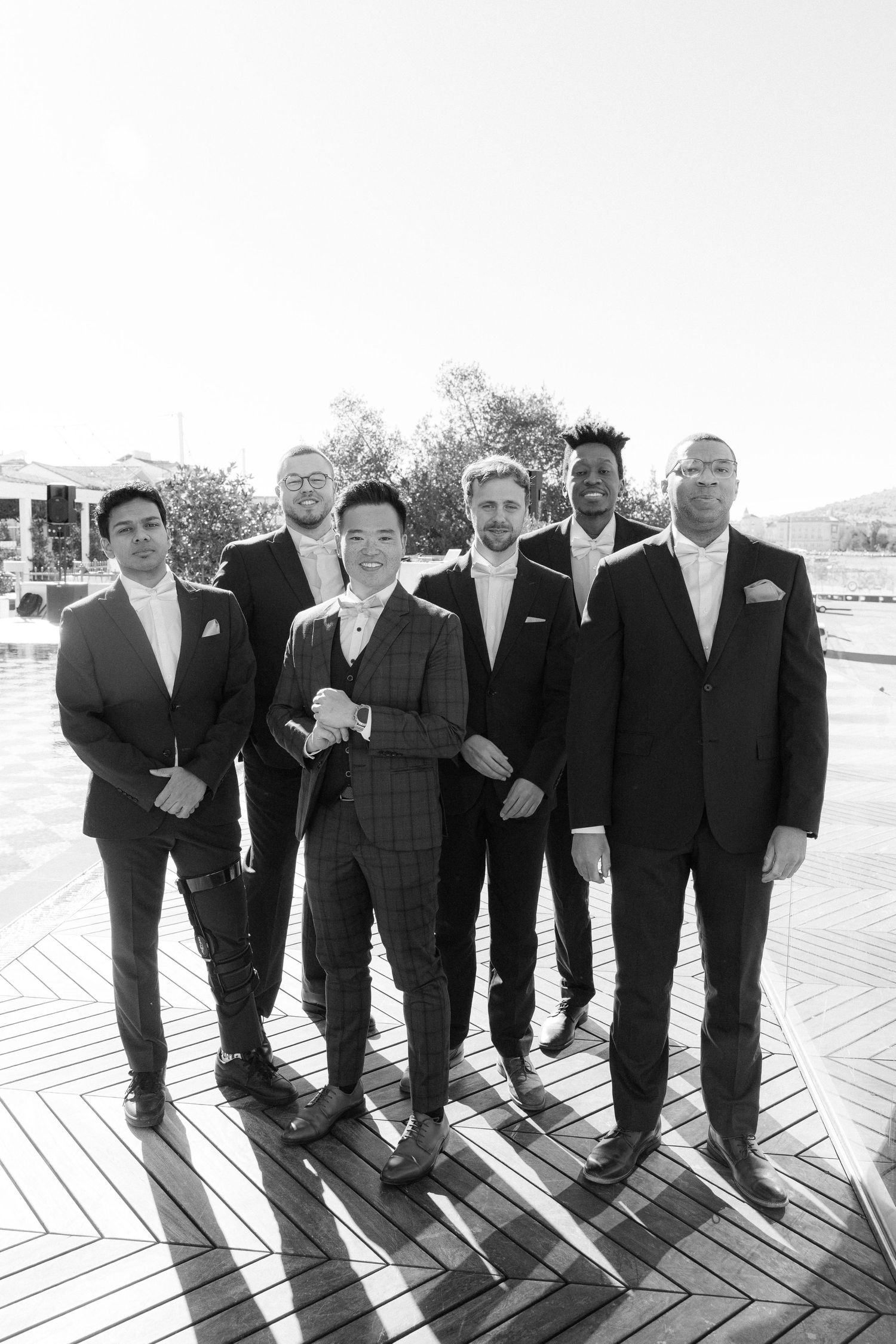 Groom in plaid three-piece suit poses with six groomsmen on outdoor deck.