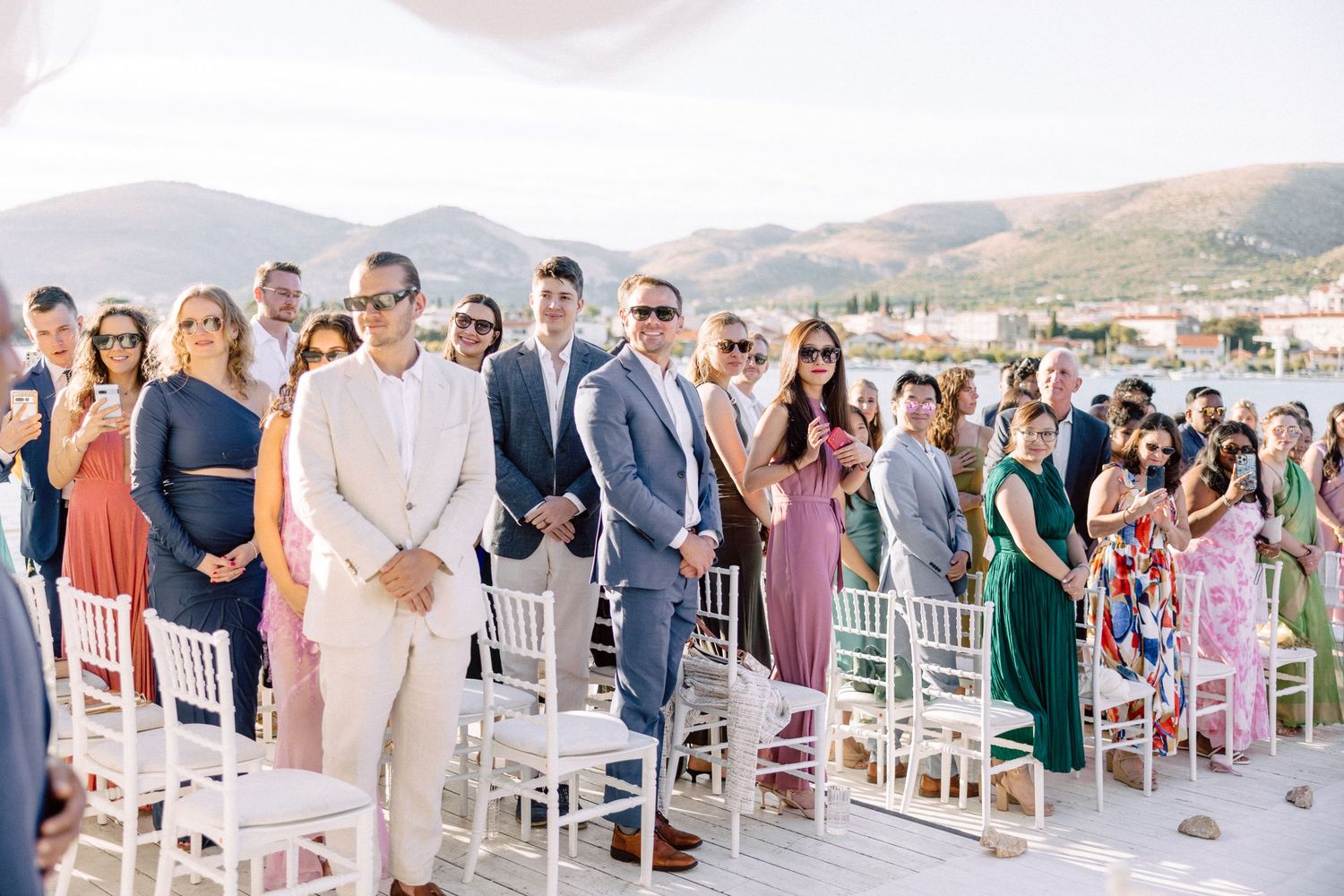 Wedding guests stand at outdoor ceremony with mountain views and white chiavari chairs.