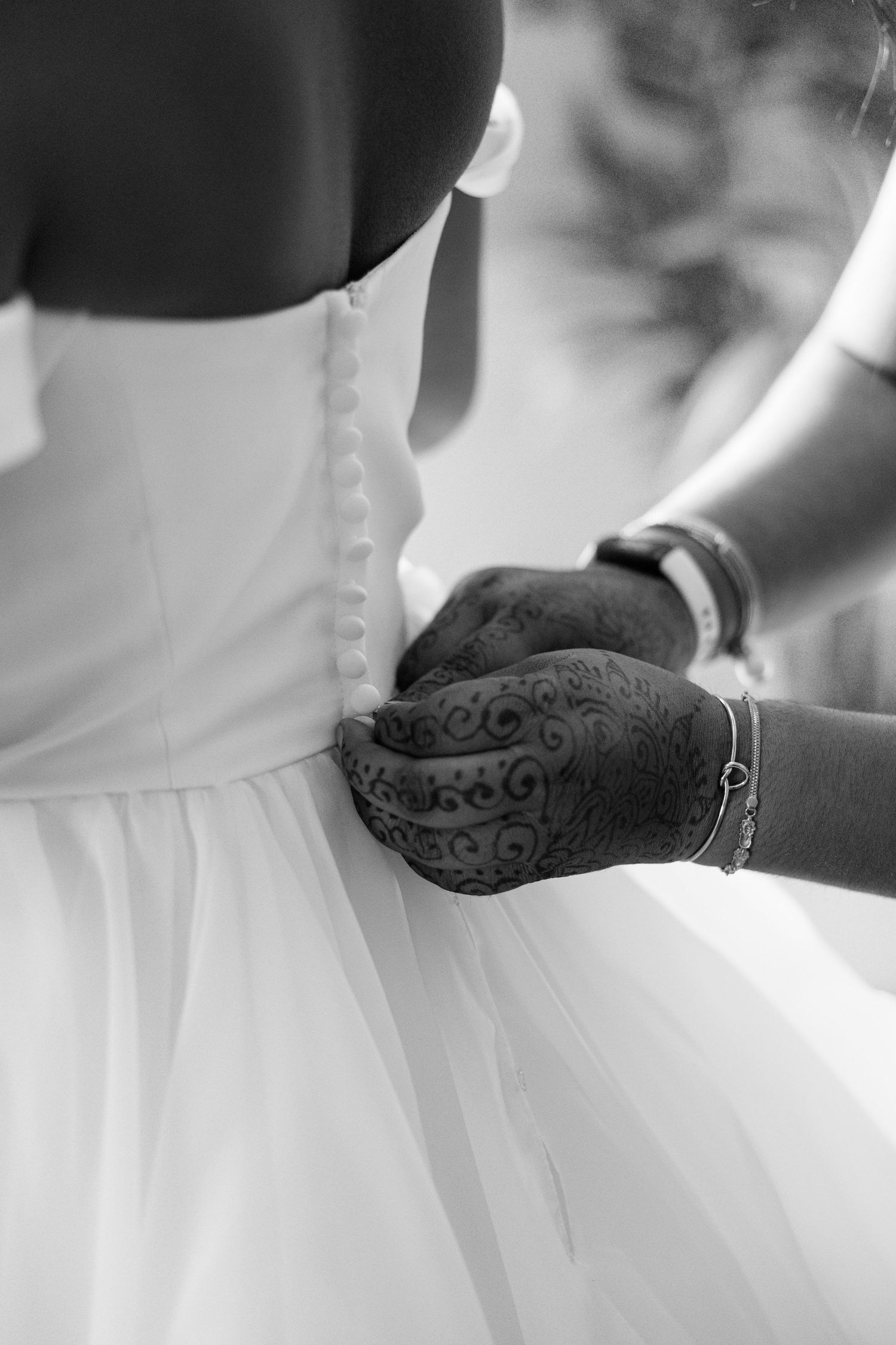 Close-up of hands with henna tattoo fastening buttons on back of bridal wedding gown in black and white.