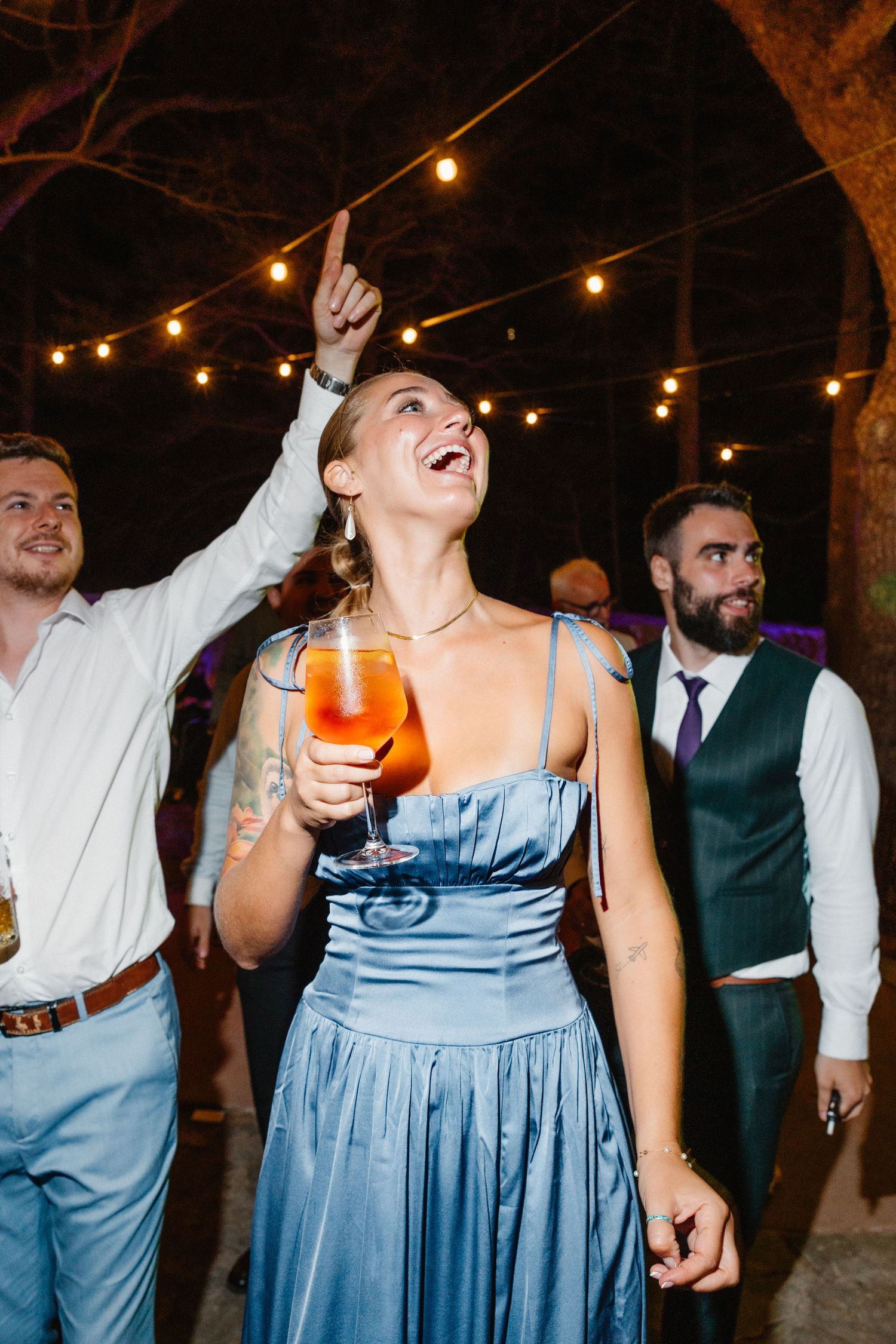 Joyful wedding guest in blue satin dress dancing with cocktail under string lights at reception.