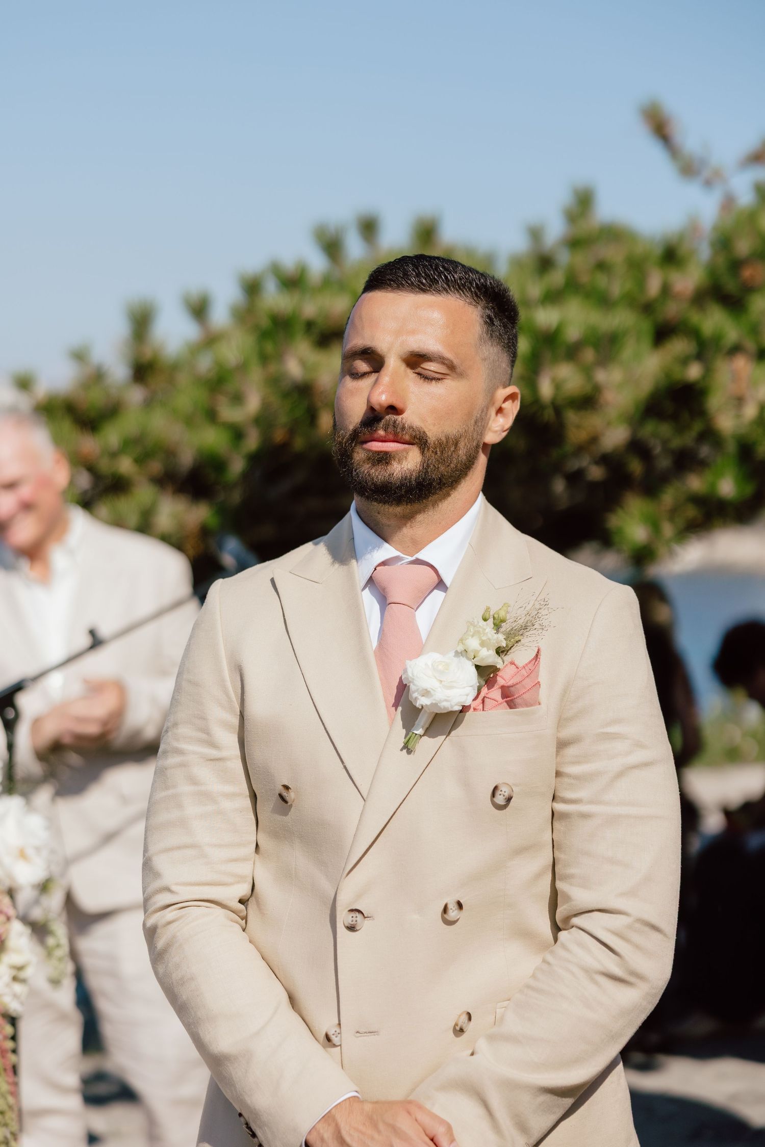 Groom in beige double-breasted suit with pink tie and boutonniere stands with eyes closed at outdoor wedding ceremony.