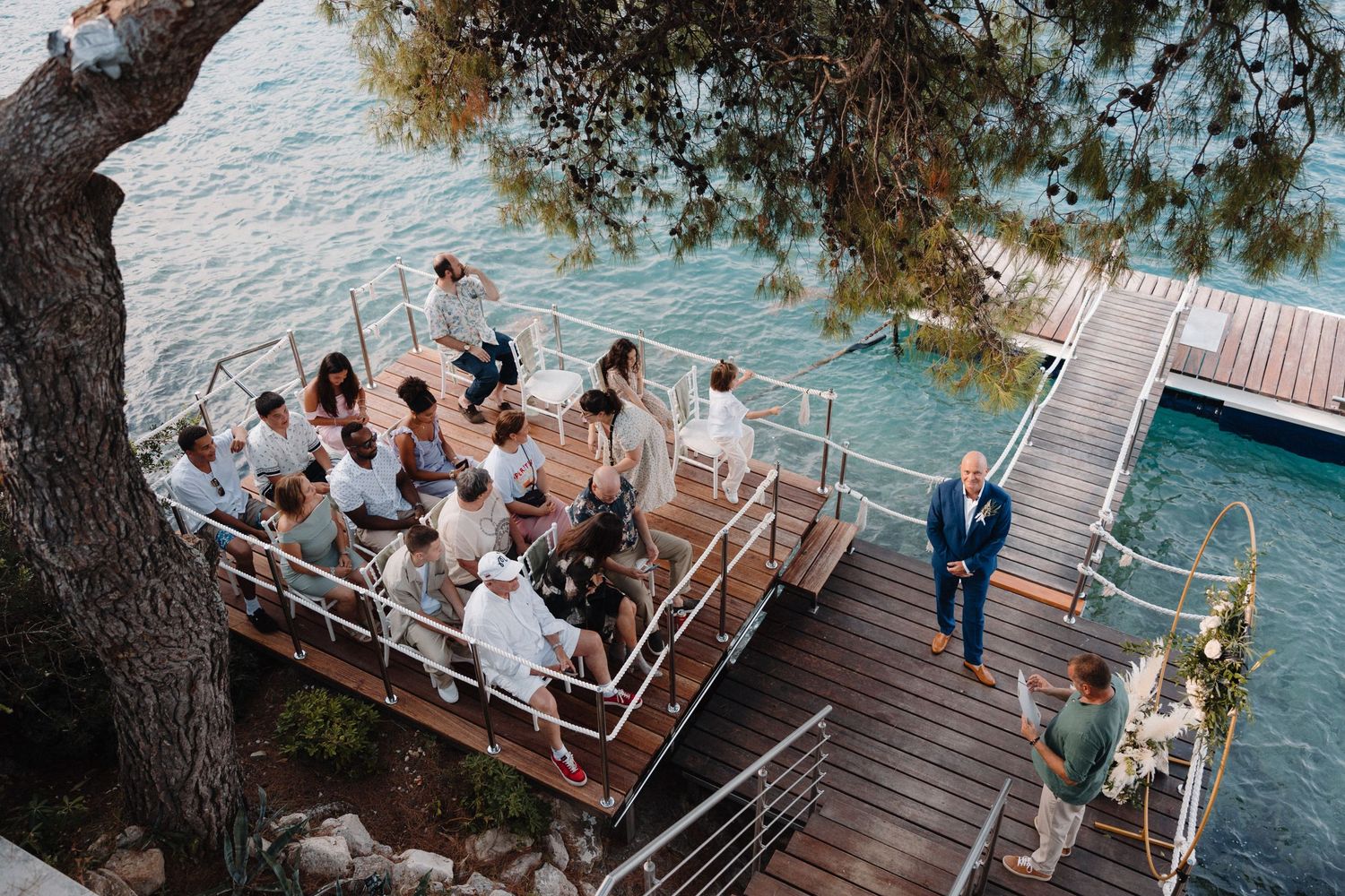 Wedding guests gather on a wooden deck overlooking turquoise waters during an outdoor ceremony.