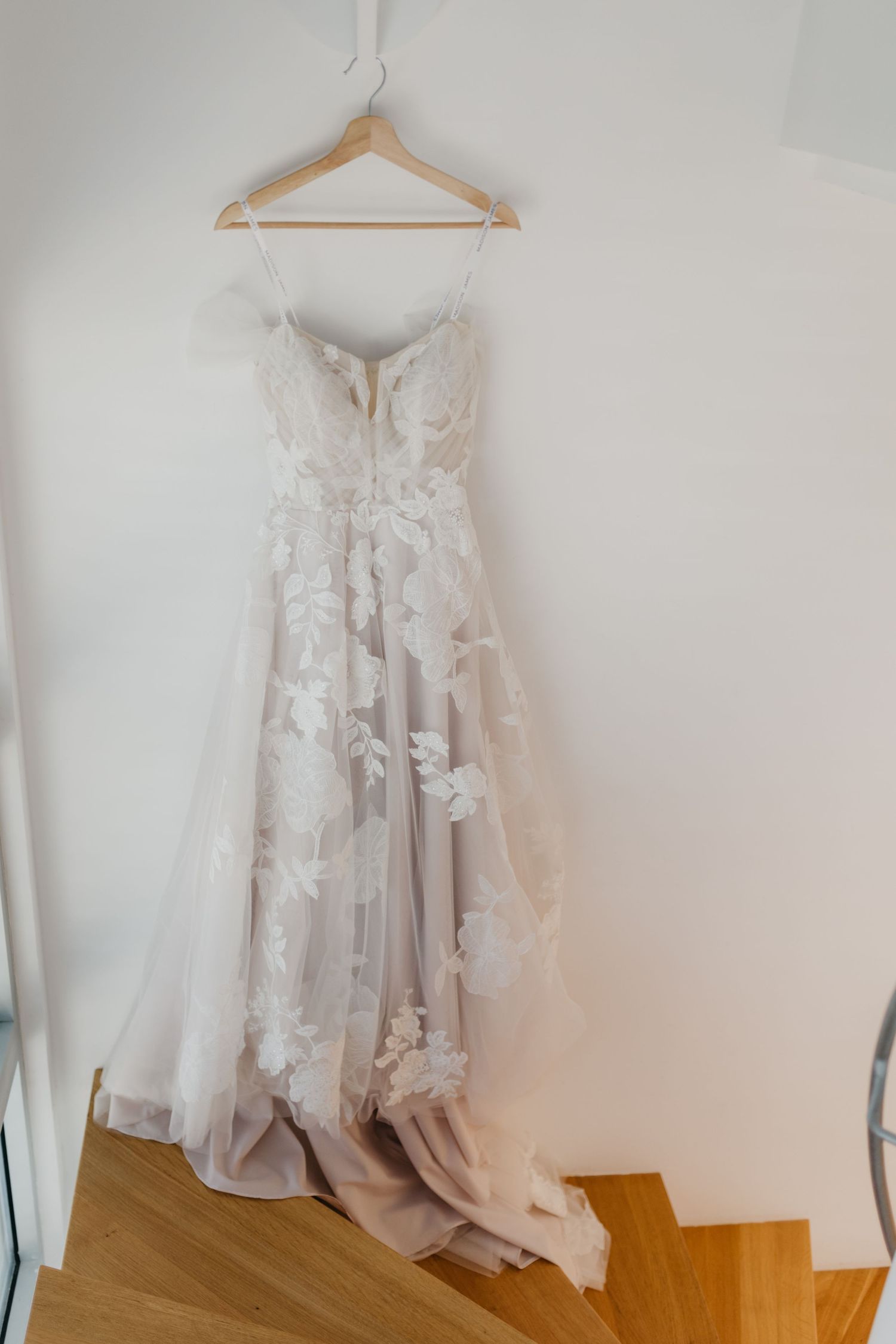 White lace bridal gown hanging on wooden hanger against white wall.