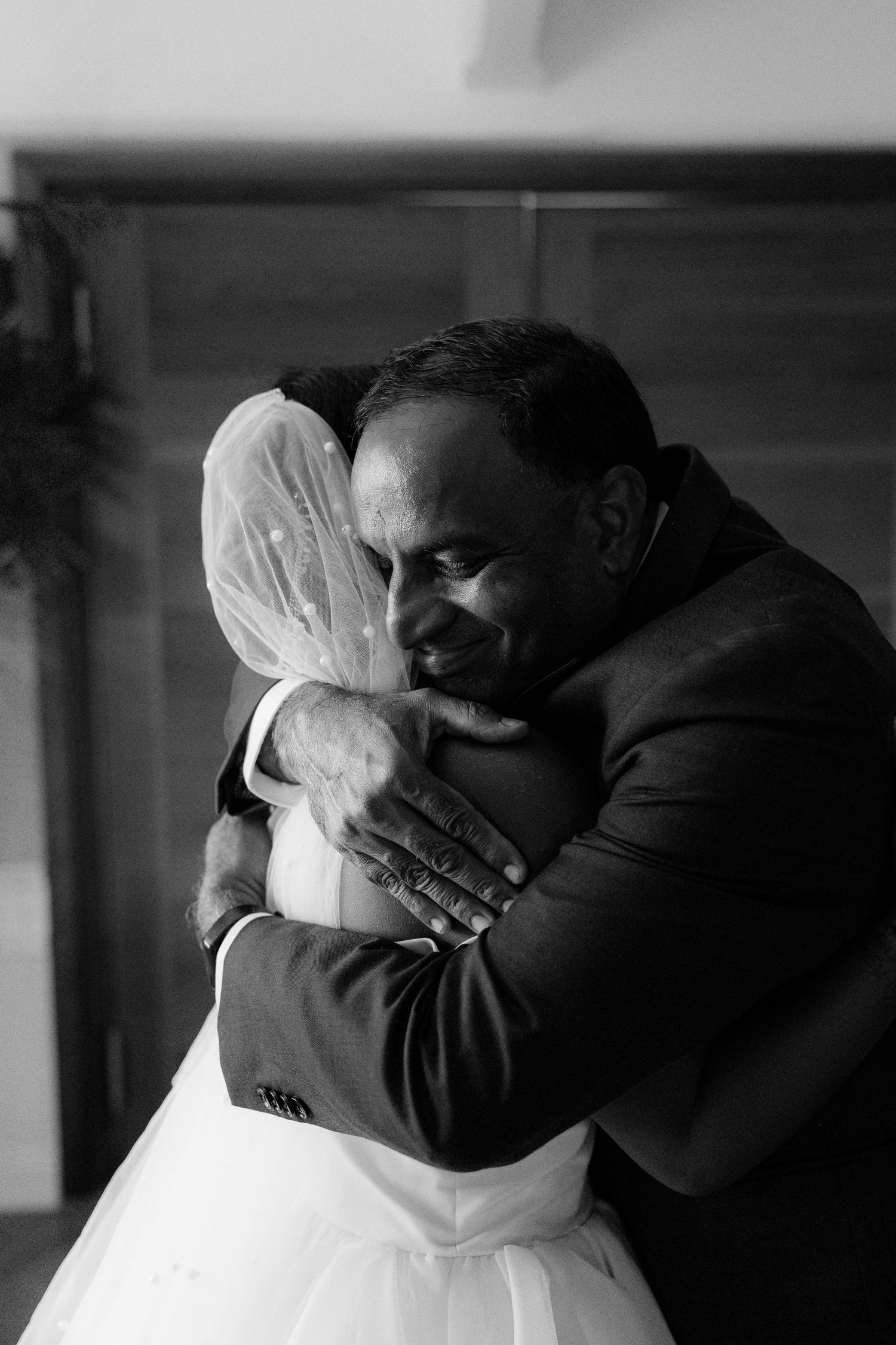 Emotional black and white photo of father embracing bride in wedding dress and veil.