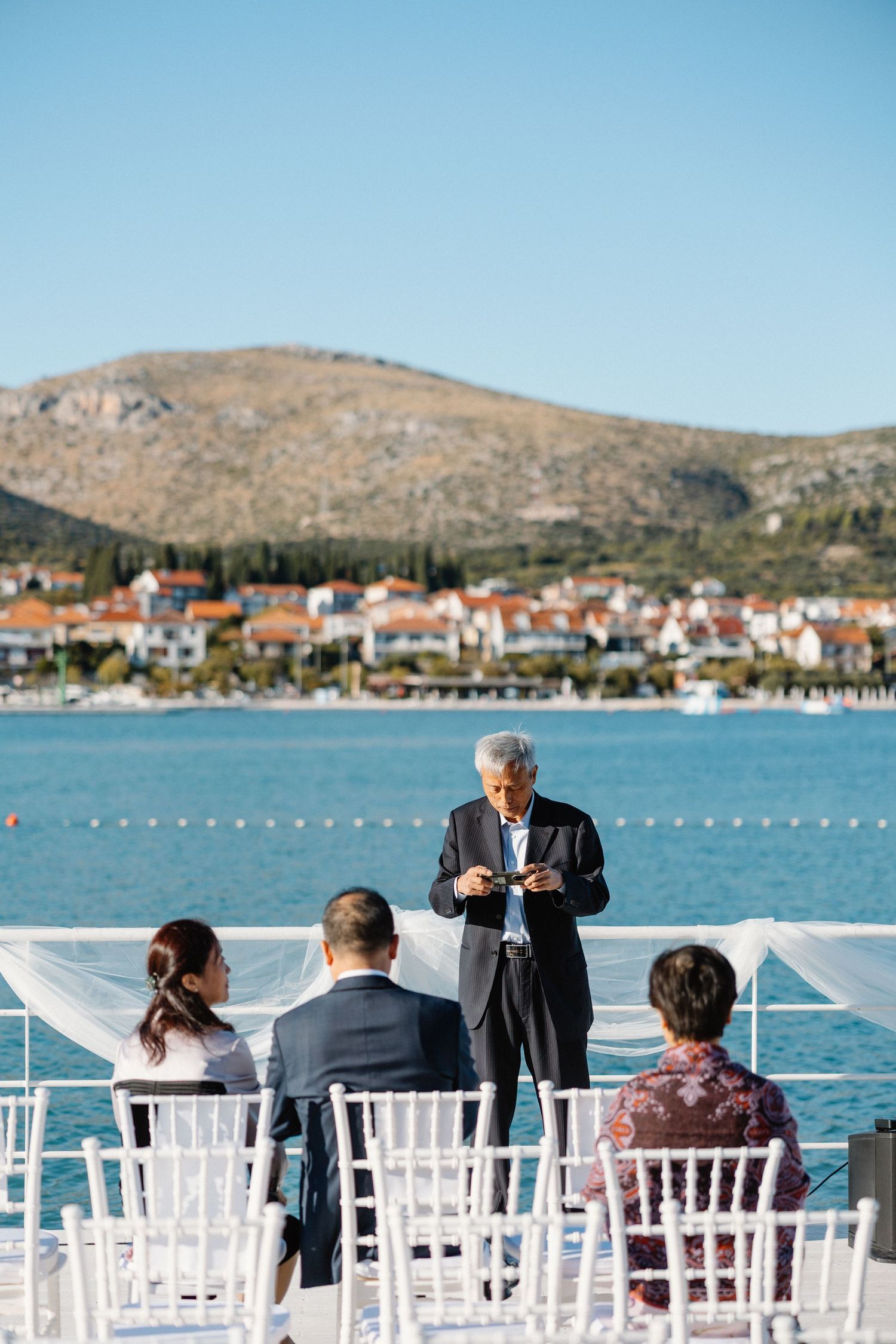 Intimate outdoor waterfront wedding ceremony with officiant and small group of guests seated in white chairs.