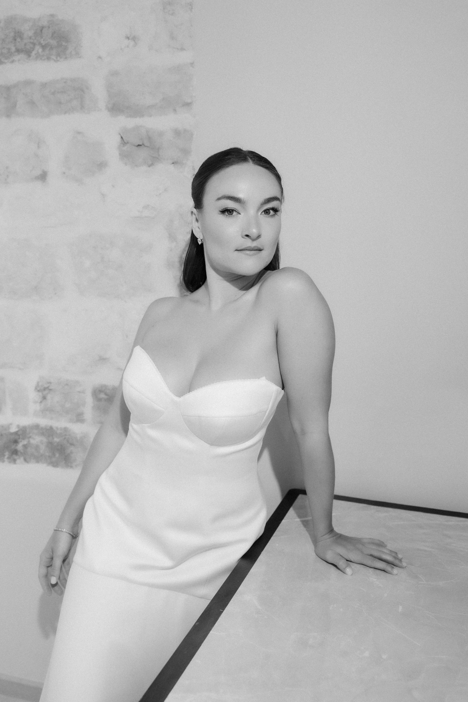 Elegant bride in strapless white satin gown posing against stone wall in black and white photo.