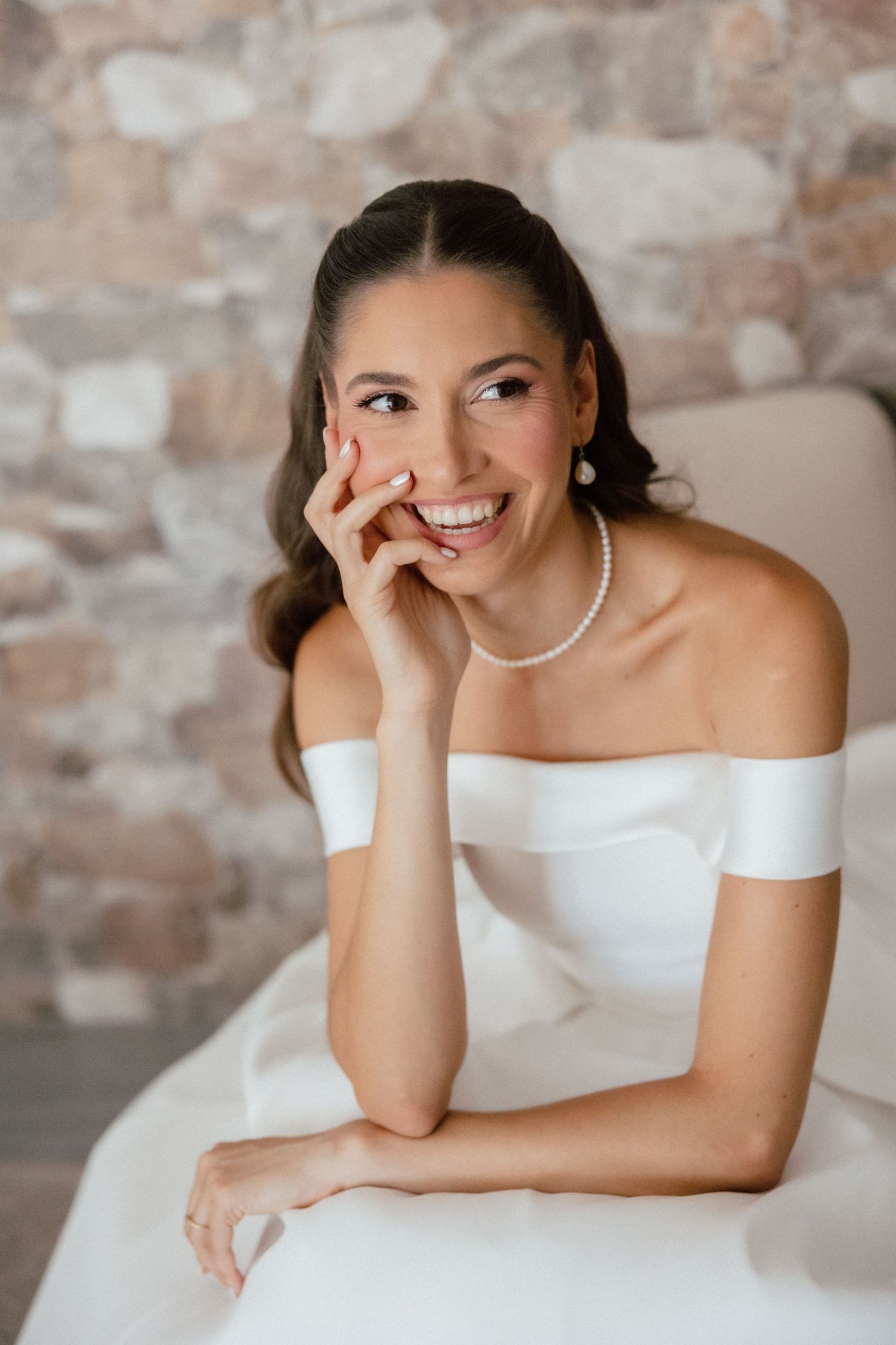 Smiling bride in off-shoulder white gown with pearl necklace and earrings, posing gracefully against a stone wall.