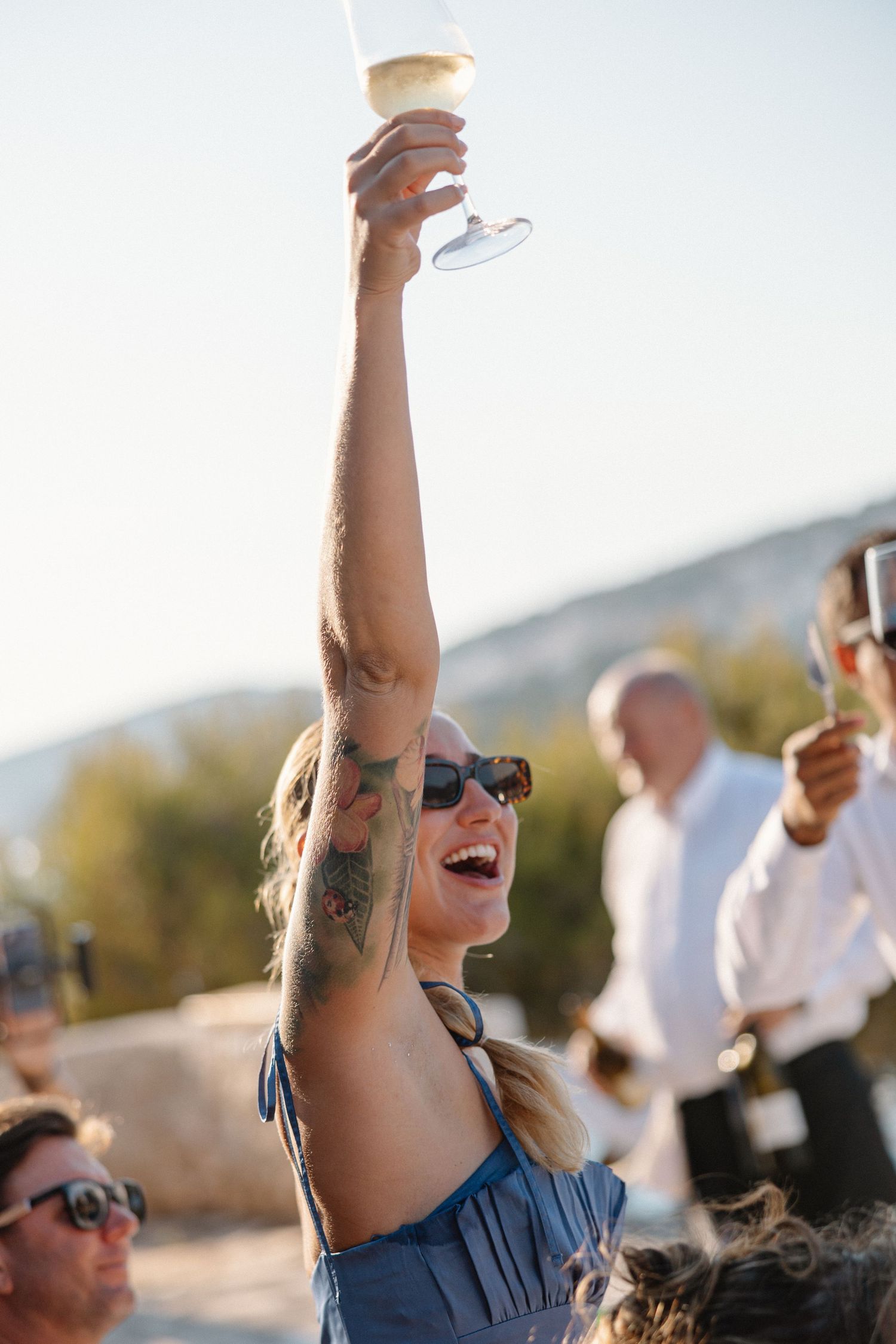 Laughing woman with tattoos raises champagne glass at outdoor wedding celebration.