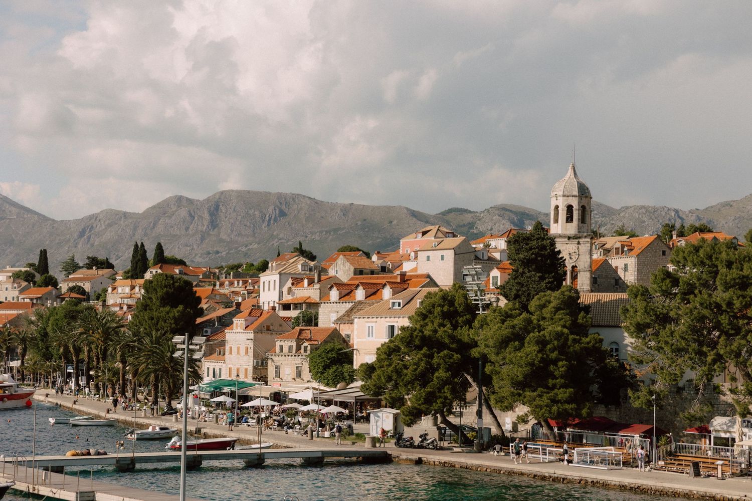 A panoramic view of a coastal Mediterranean town with church steeples and mountains in the background.