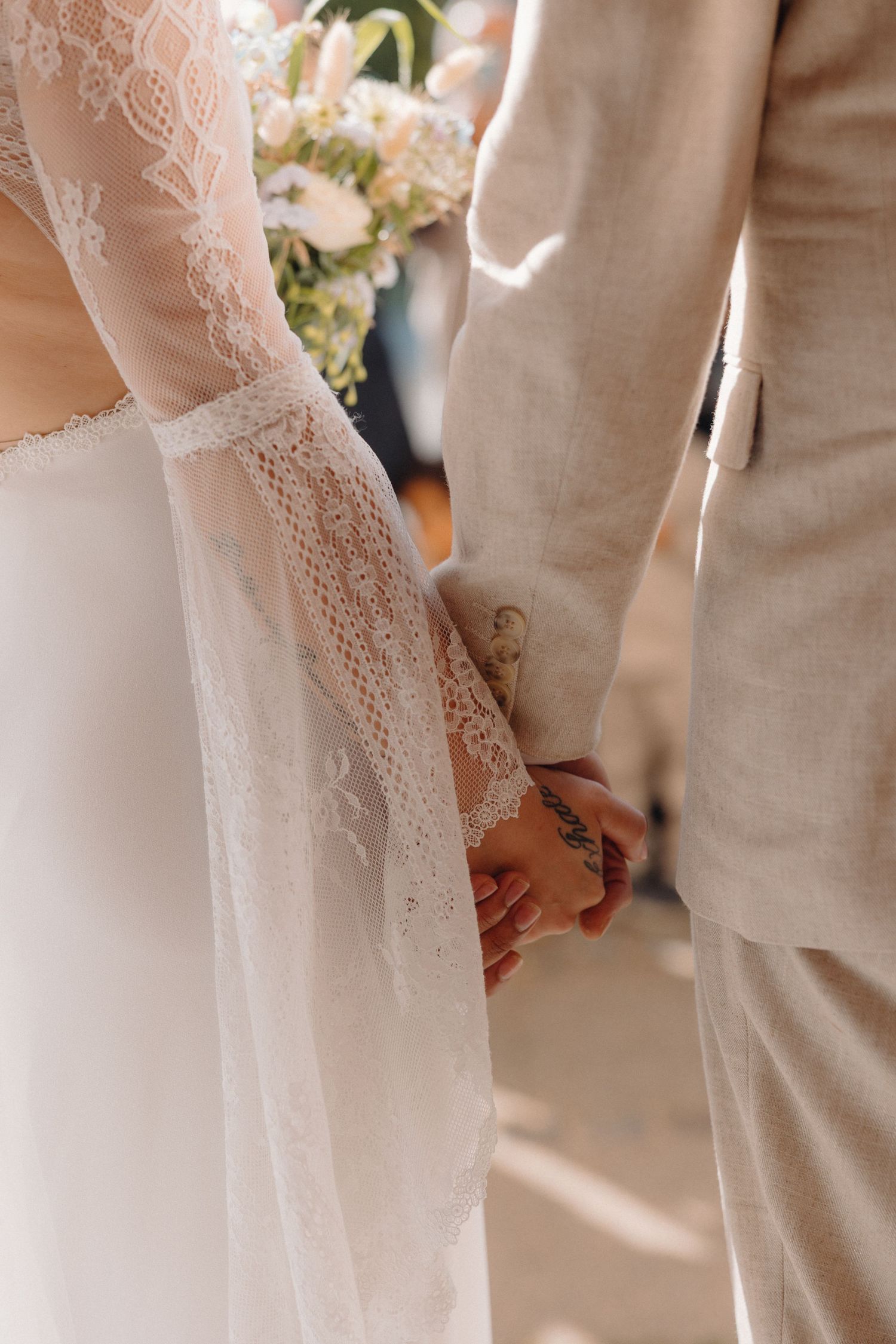 Detail shot of bride and groom holding hands, with lace wedding dress sleeve and beige suit visible.