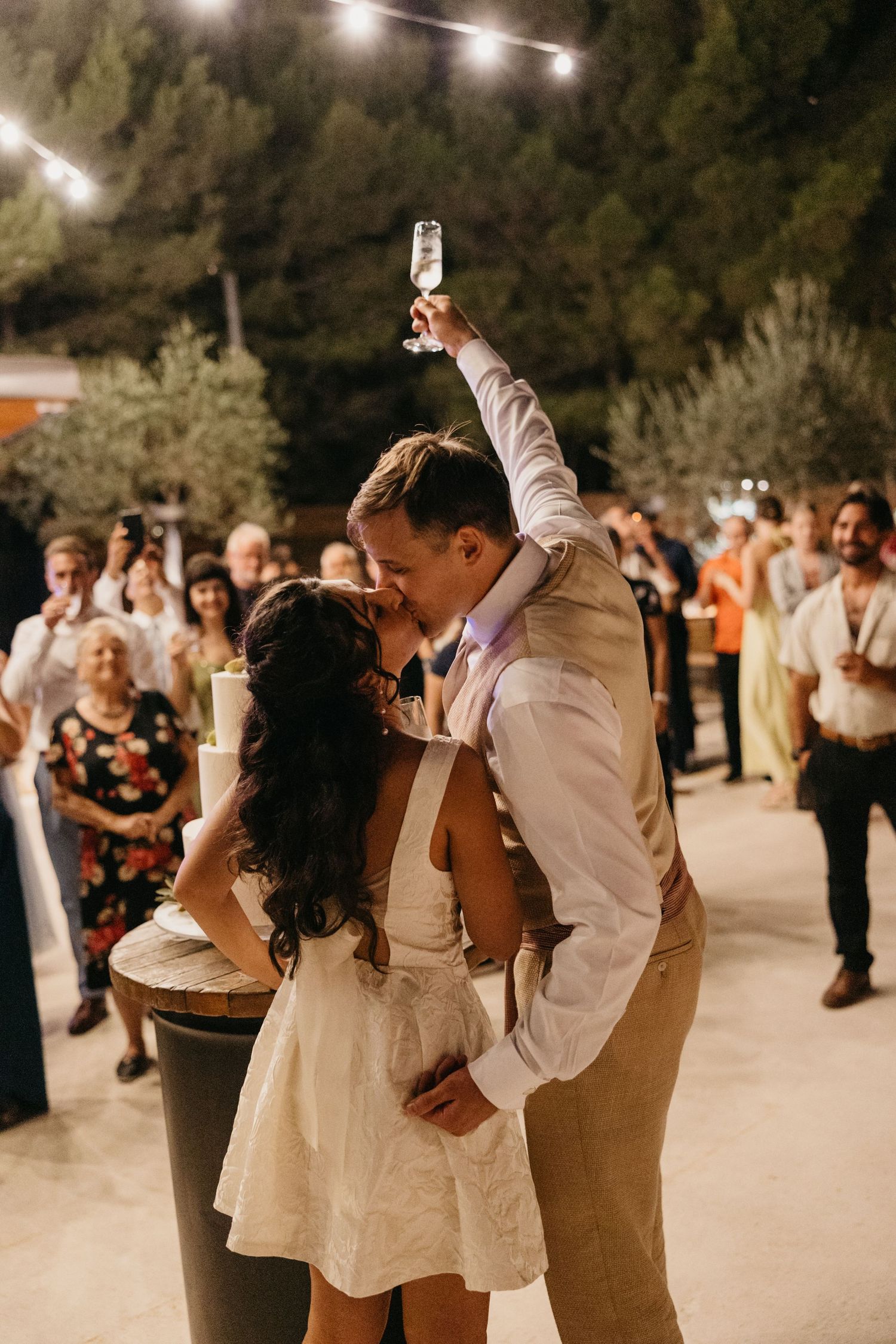 Couple dancing at outdoor wedding reception under string lights with guests watching.