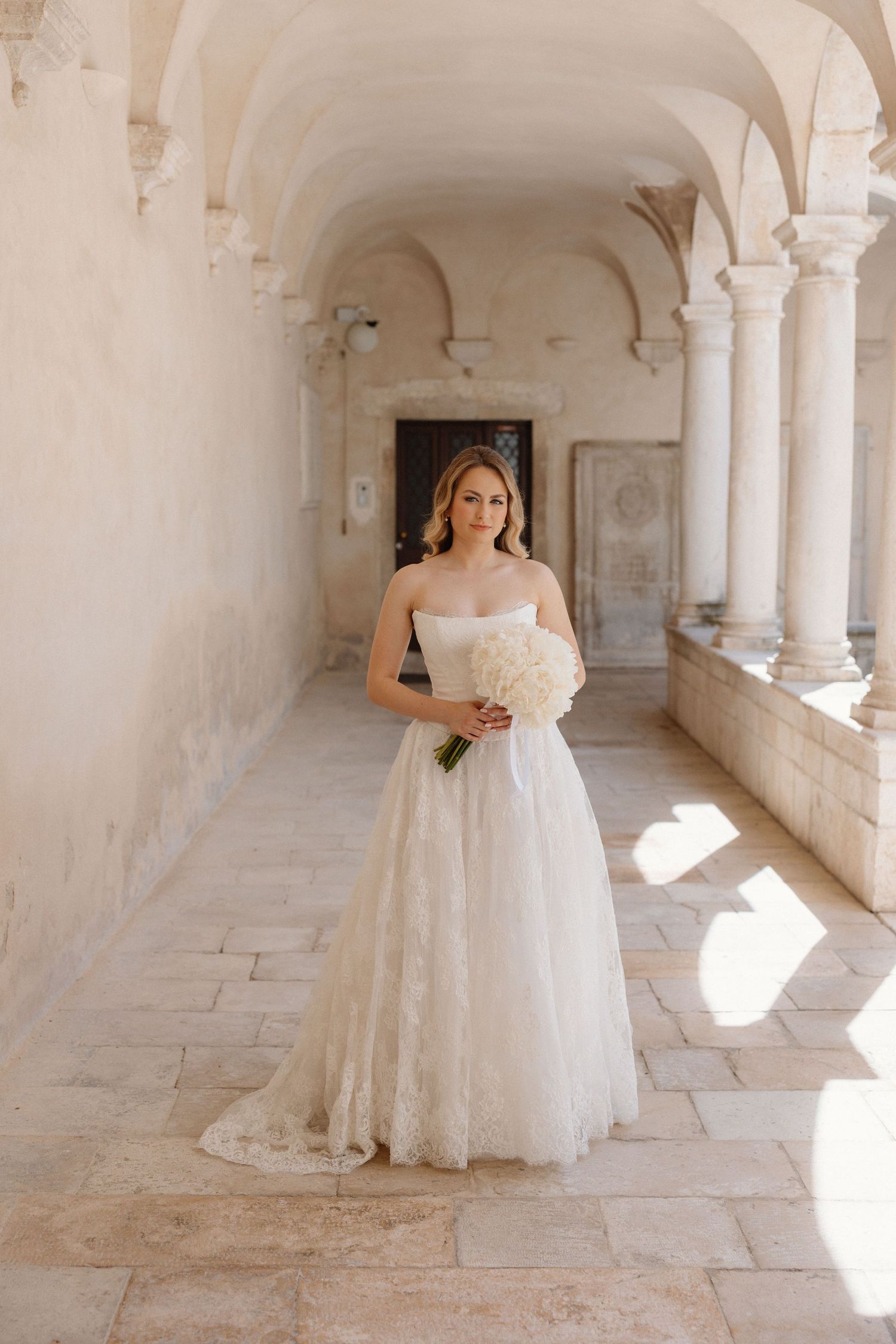 A bride in an elegant white lace gown stands in a historic stone corridor with arched columns and natural sunlight.