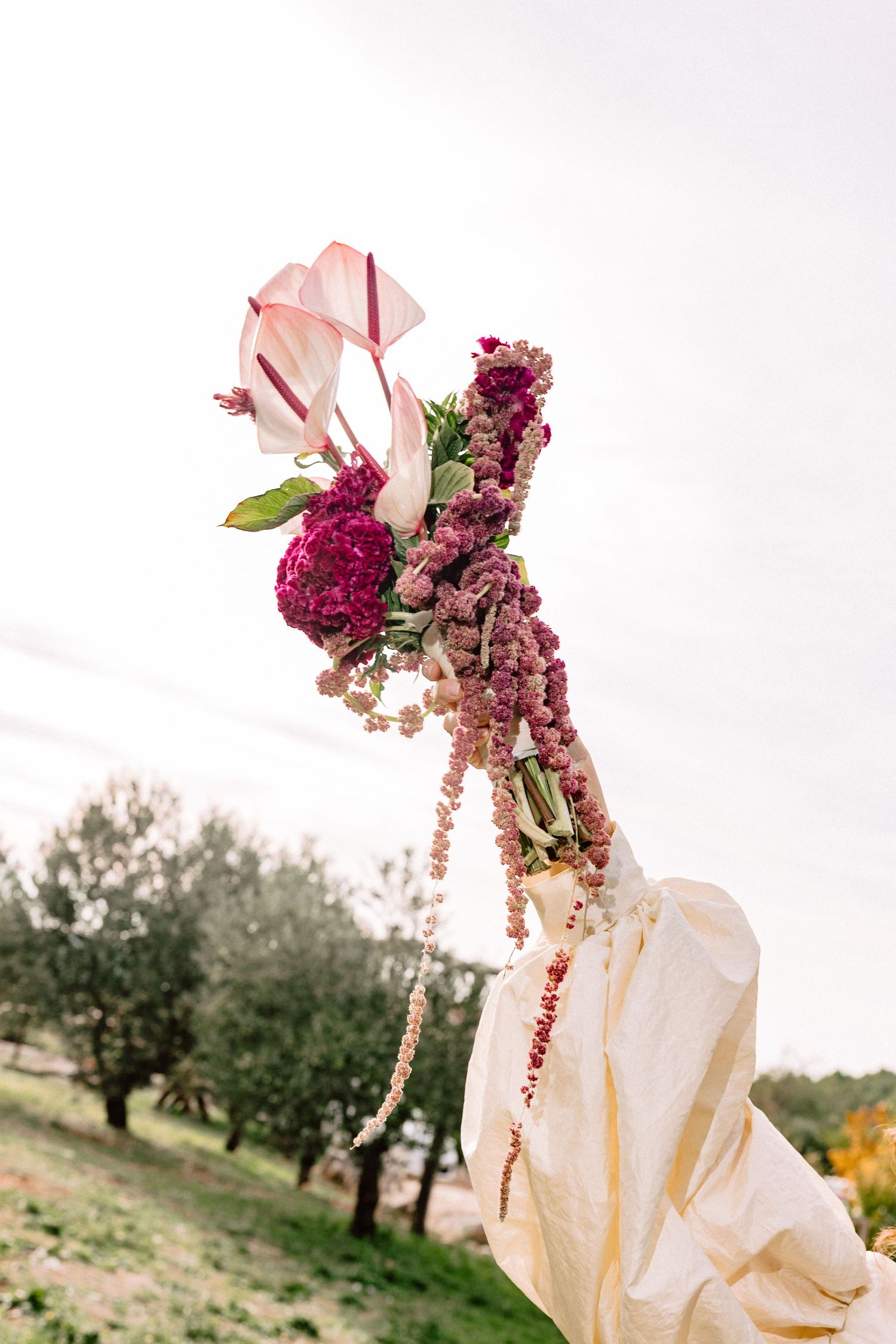 Pink and purple floral bouquet against white sky with olive trees in background.
