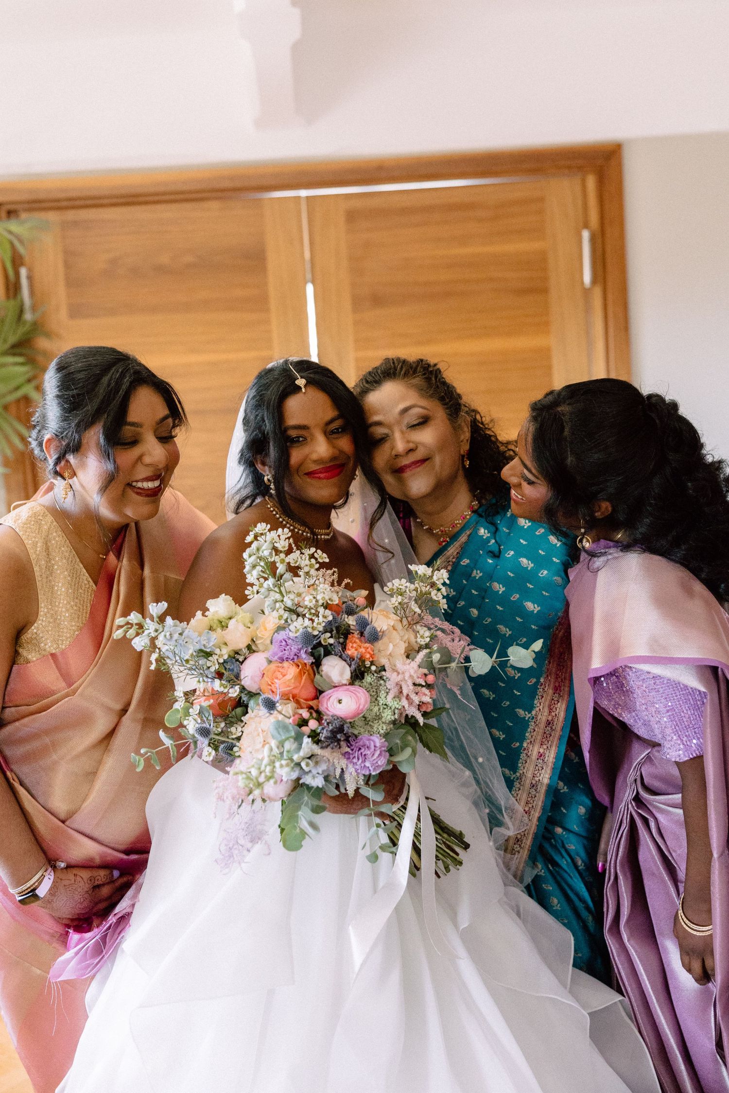 A radiant bride in a white gown holds a colorful bouquet surrounded by three women in vibrant Indian sarees.