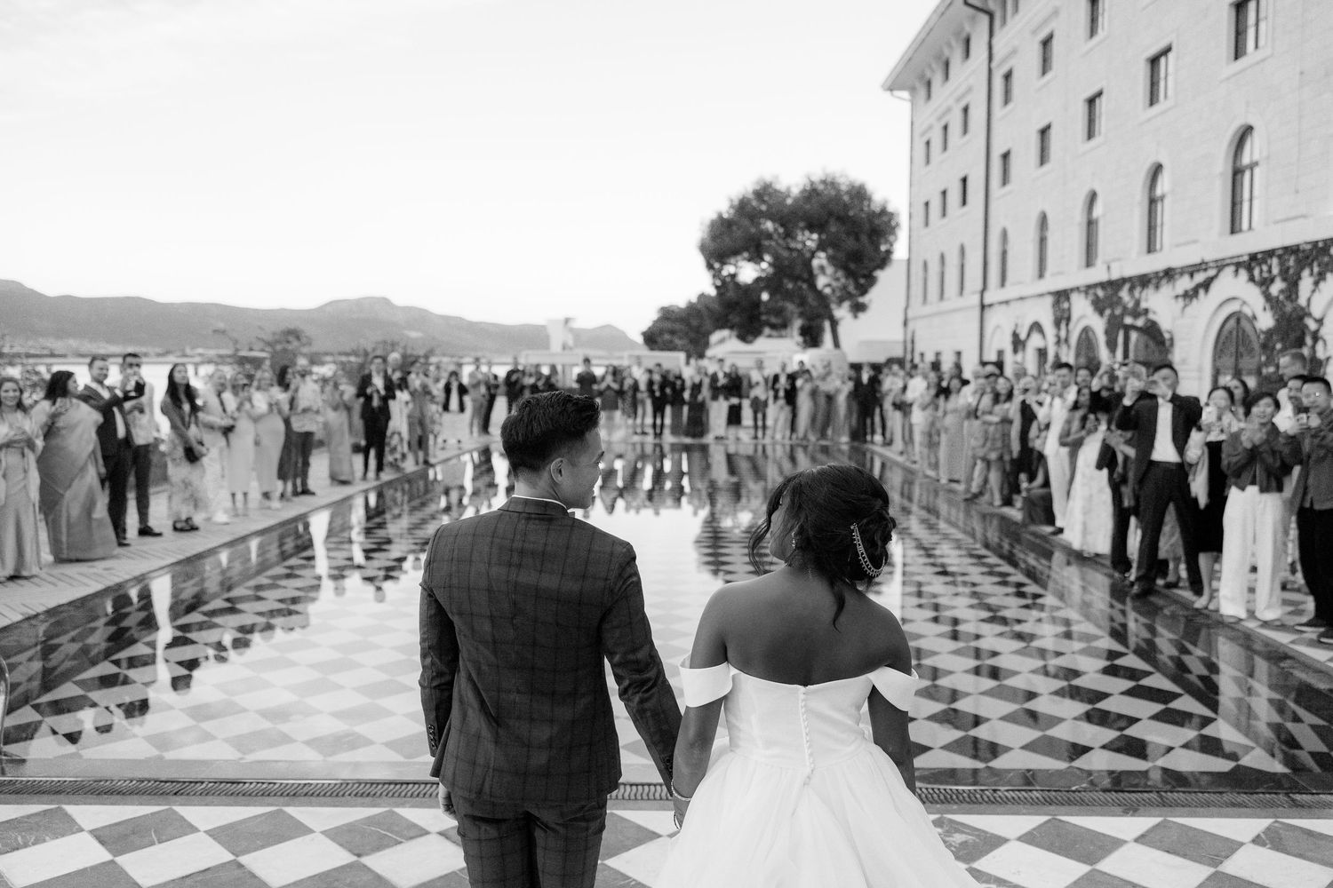 Bride and groom walk hand-in-hand toward cheering guests on a grand checkered courtyard in B&W.