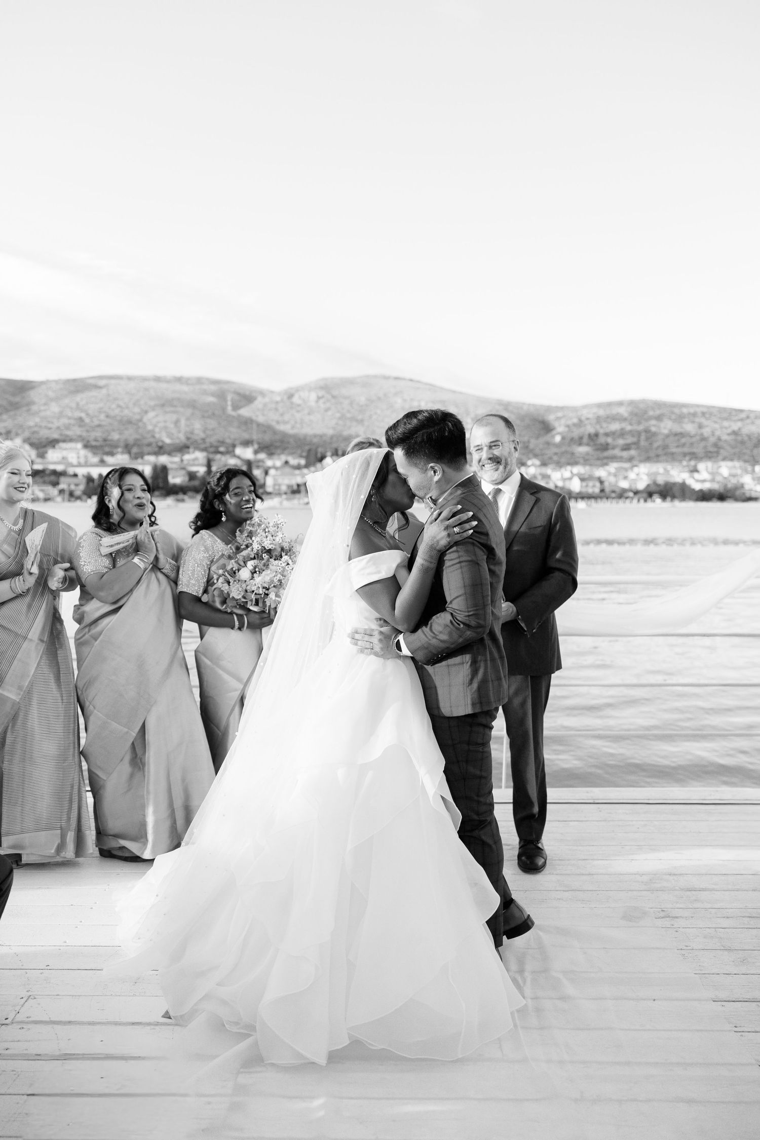 Bride and groom share first kiss on waterfront dock with joyful wedding party celebrating behind them.