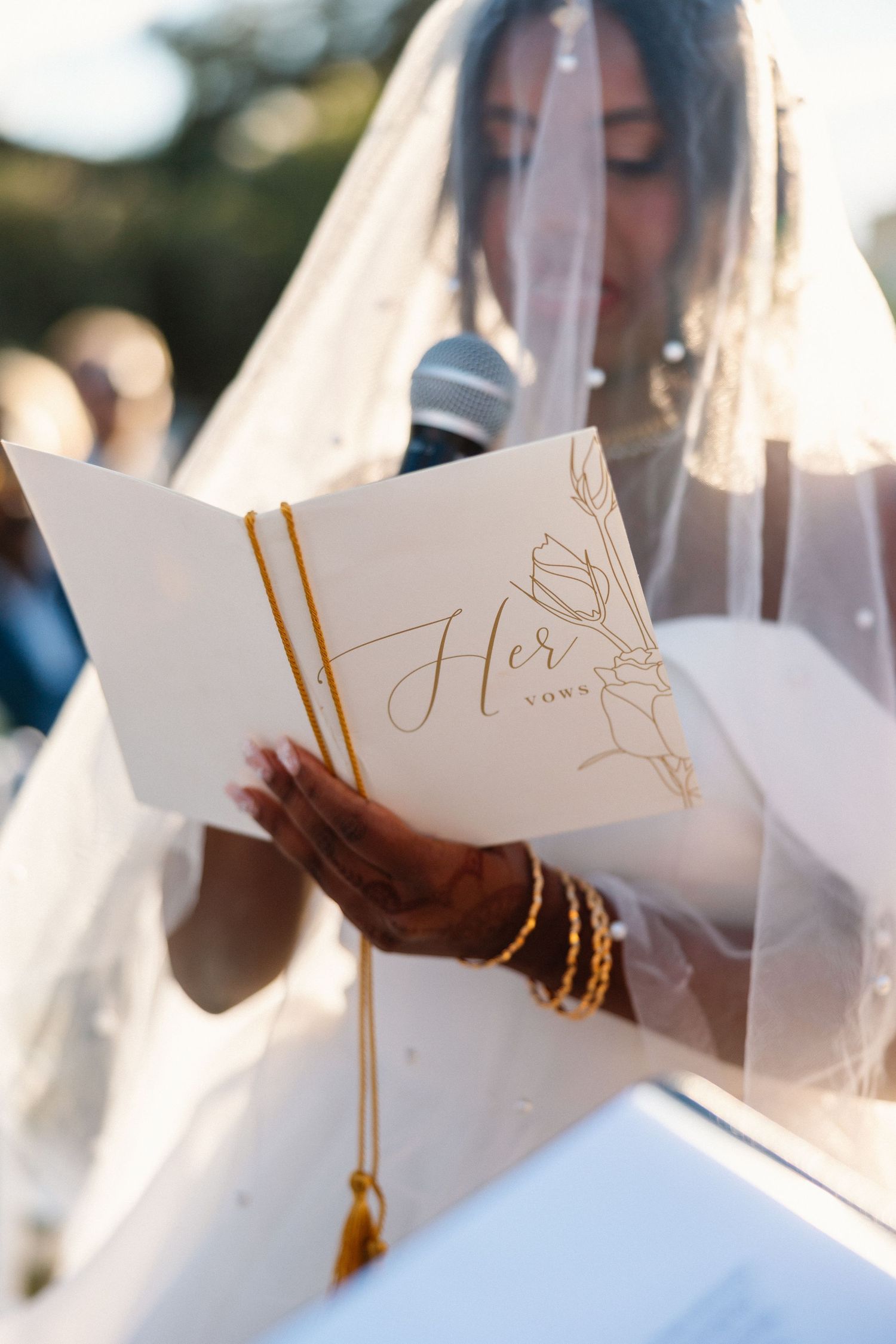 Bride in white veil reads from elegant gold-lettered Her Vows booklet during outdoor wedding ceremony.