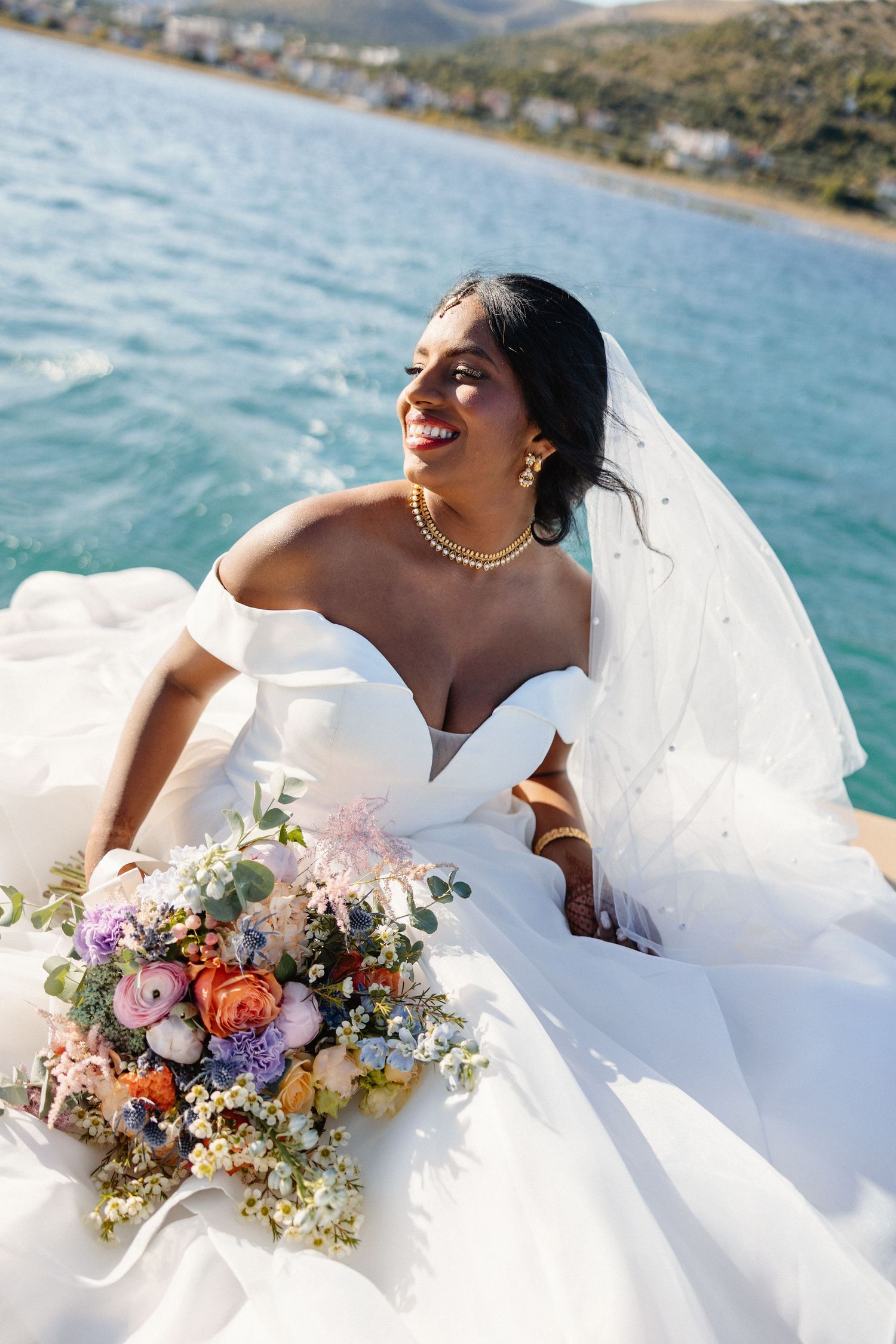 Radiant bride in white off-shoulder gown sitting on boat bow with colorful bouquet by the sea.