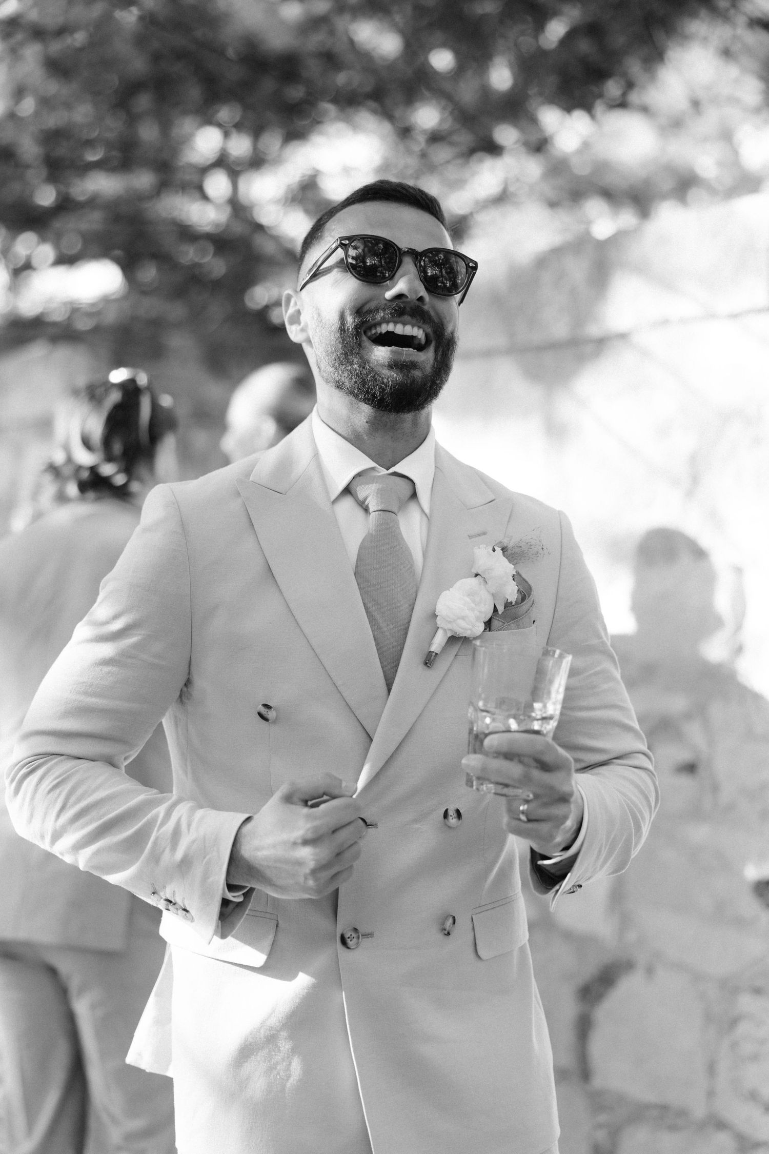 Dapper groom in beige double-breasted suit with boutonniere and sunglasses laughing outdoors.