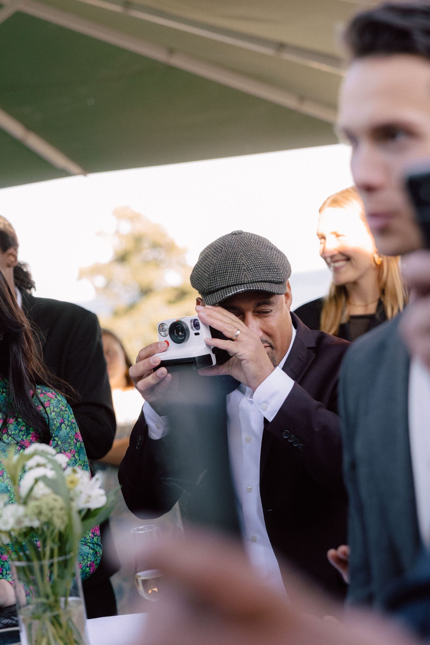 Wedding guests take photos during outdoor reception under white tent canopy.