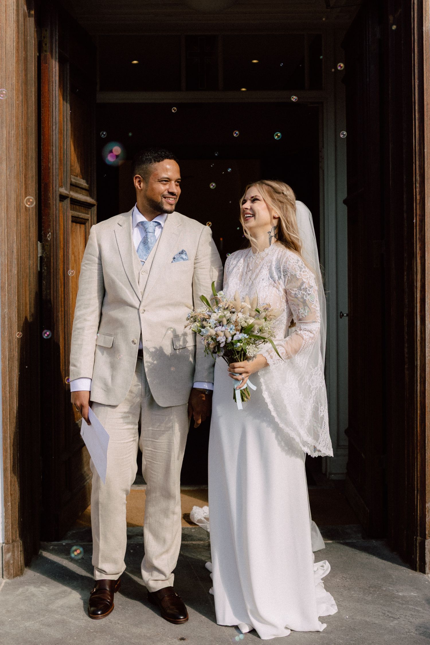 A joyful wedding couple shares a laugh while standing in a doorway surrounded by bubbles.
