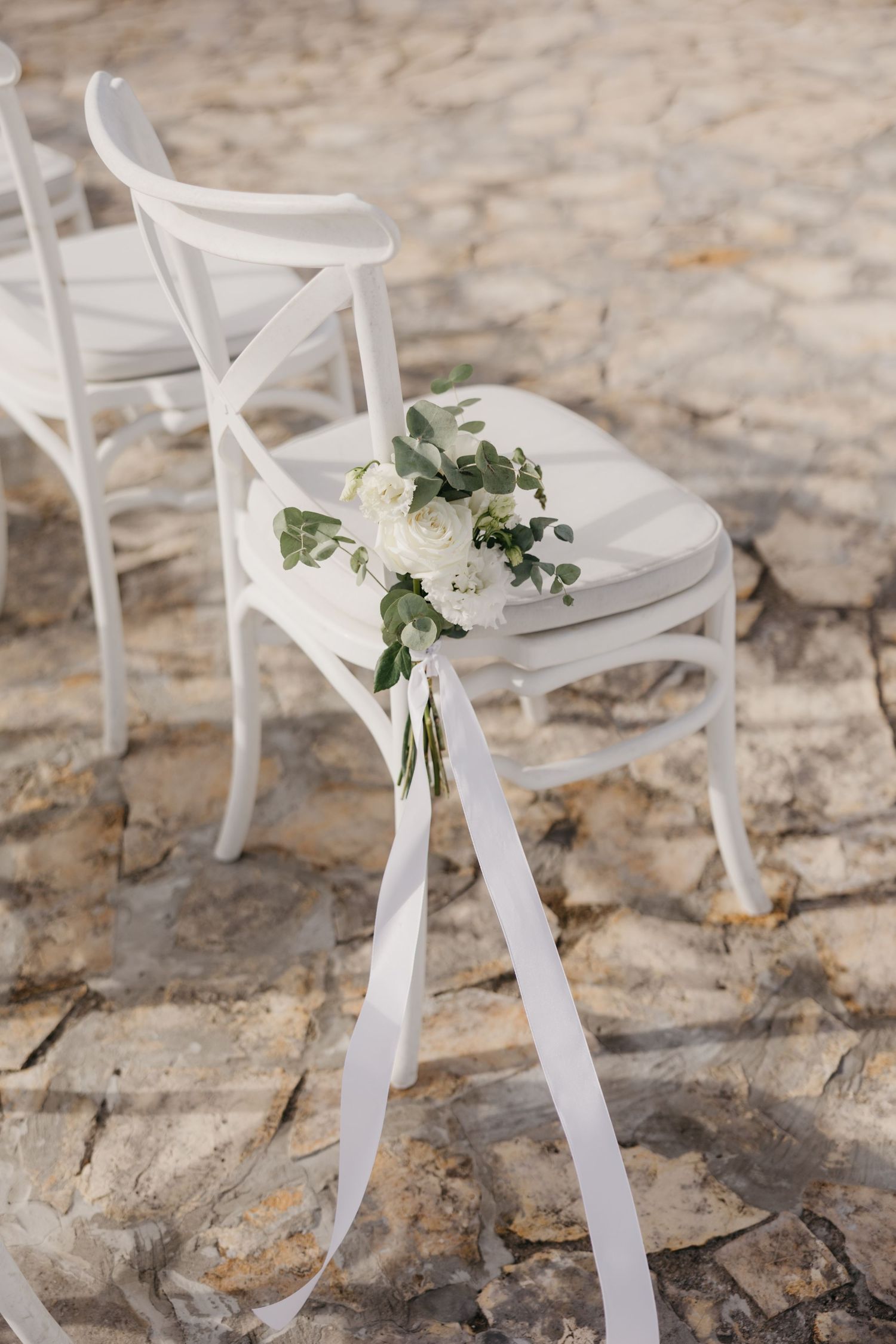White ceremony chairs decorated with small floral bouquets and ribbons on stone ground.