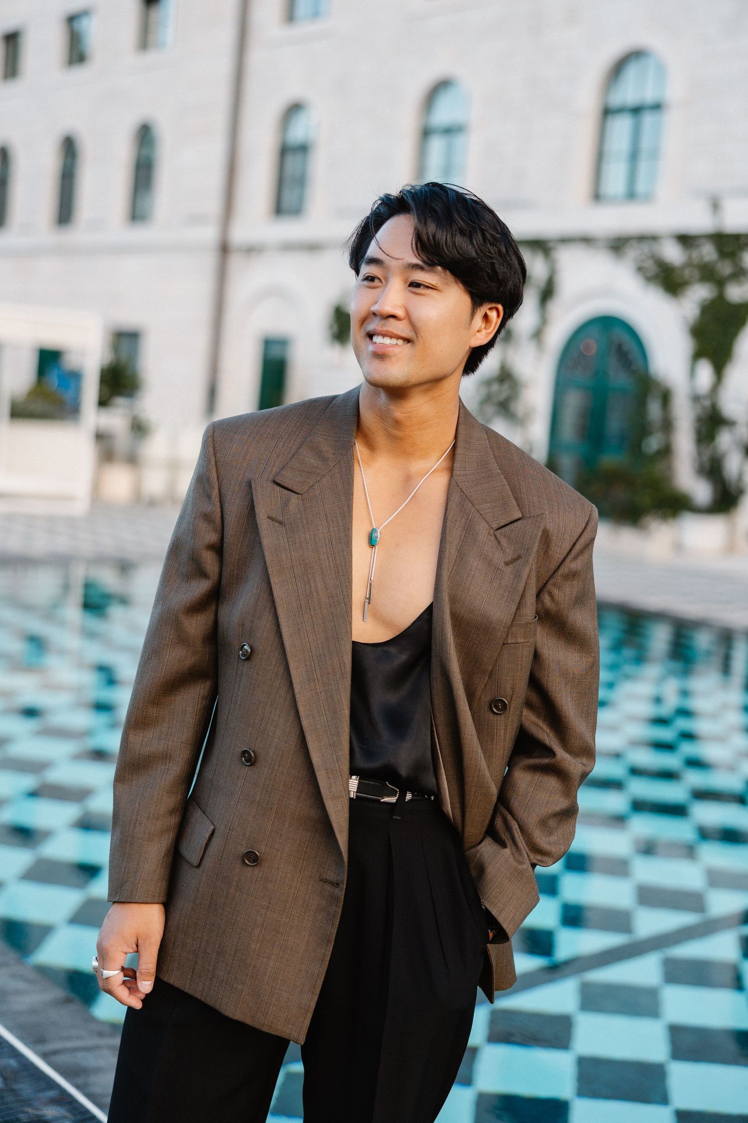 Stylish Asian man in brown blazer and black satin top smiling outdoors near a reflective pool.