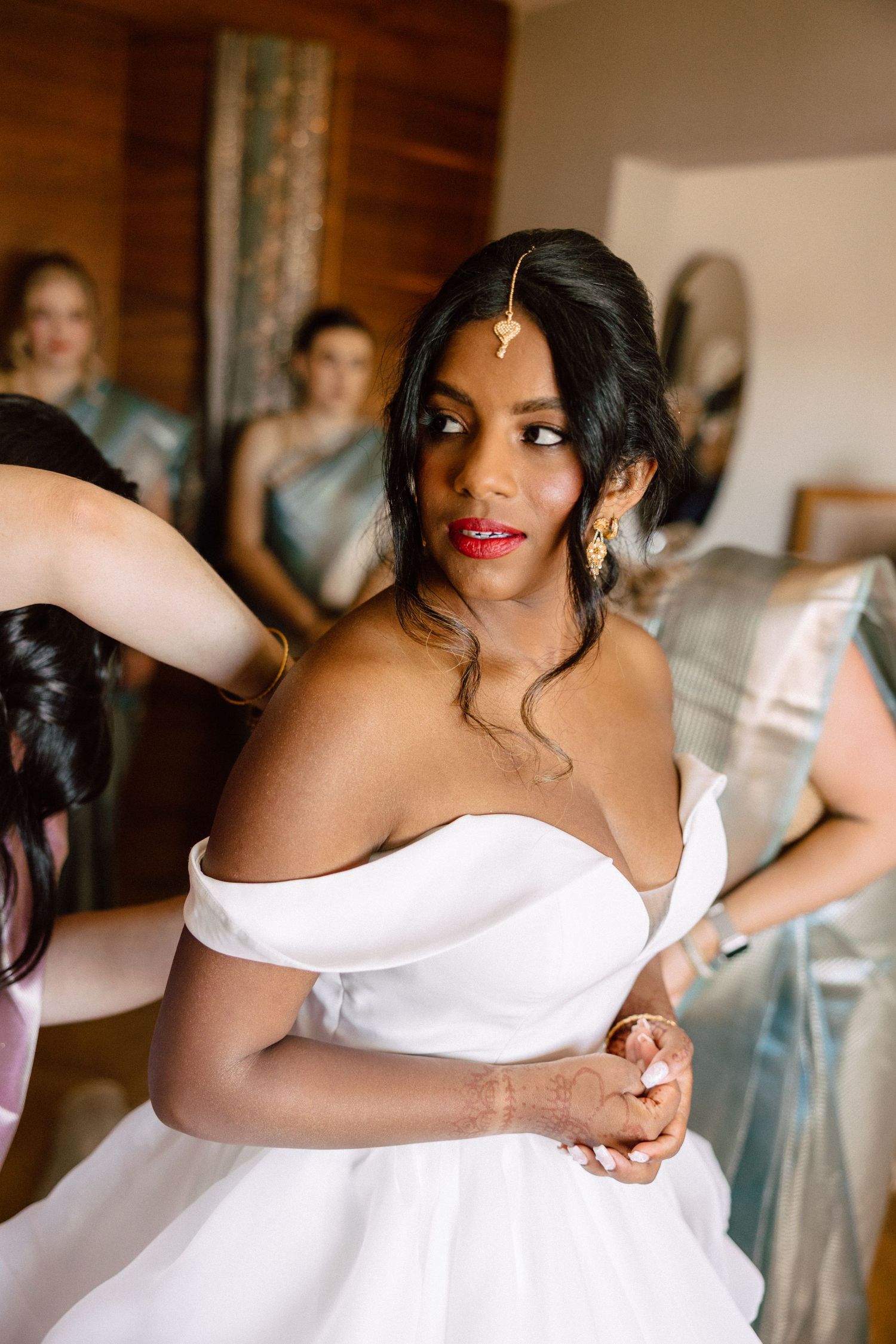 Beautiful South Asian bride in a white off-shoulder wedding gown wearing gold maang tikka and earrings getting ready.