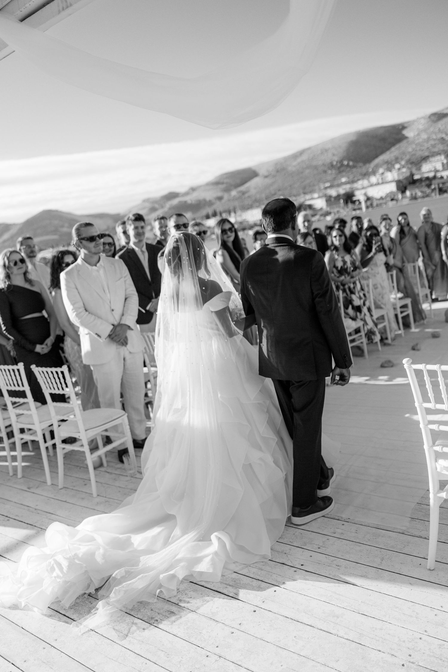 Bride in flowing gown with veil walks down aisle with groom at outdoor ceremony.