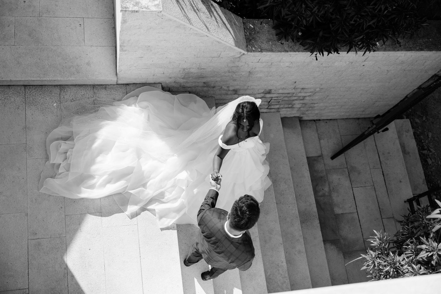 Aerial view of bride in flowing white gown descending stone stairs with companion.