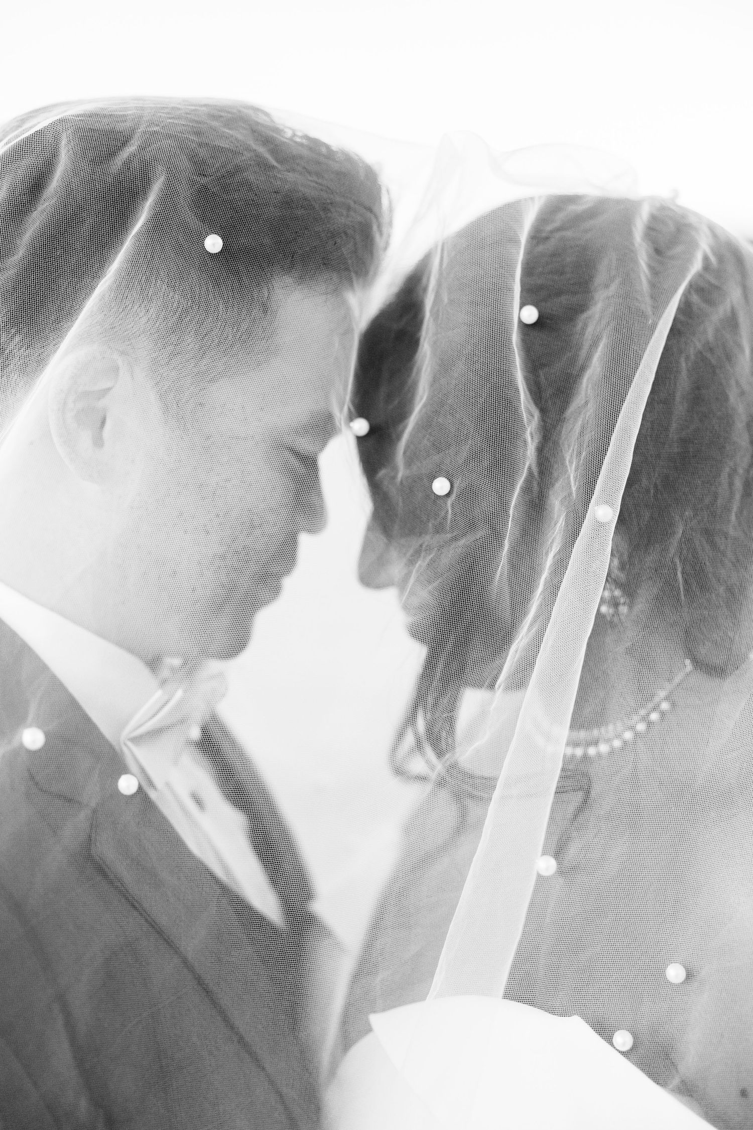 Bride and groom share intimate moment under pearl-adorned veil in soft black and white portrait.