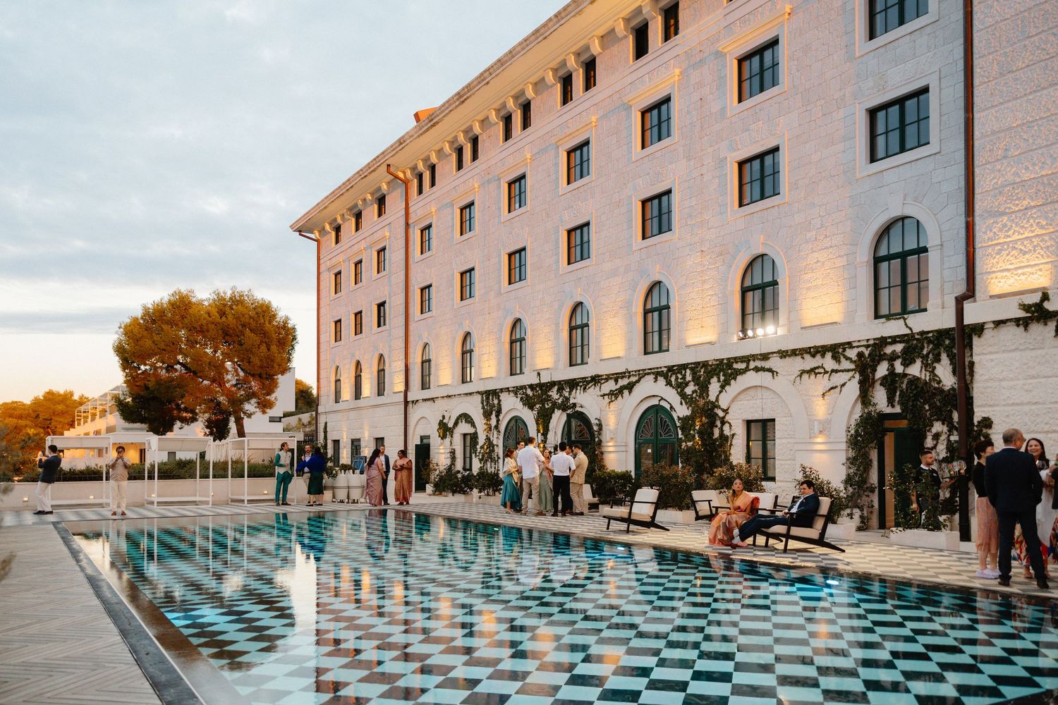 Guests mingle beside illuminated checkered pool at luxury hotel during golden hour wedding reception.
