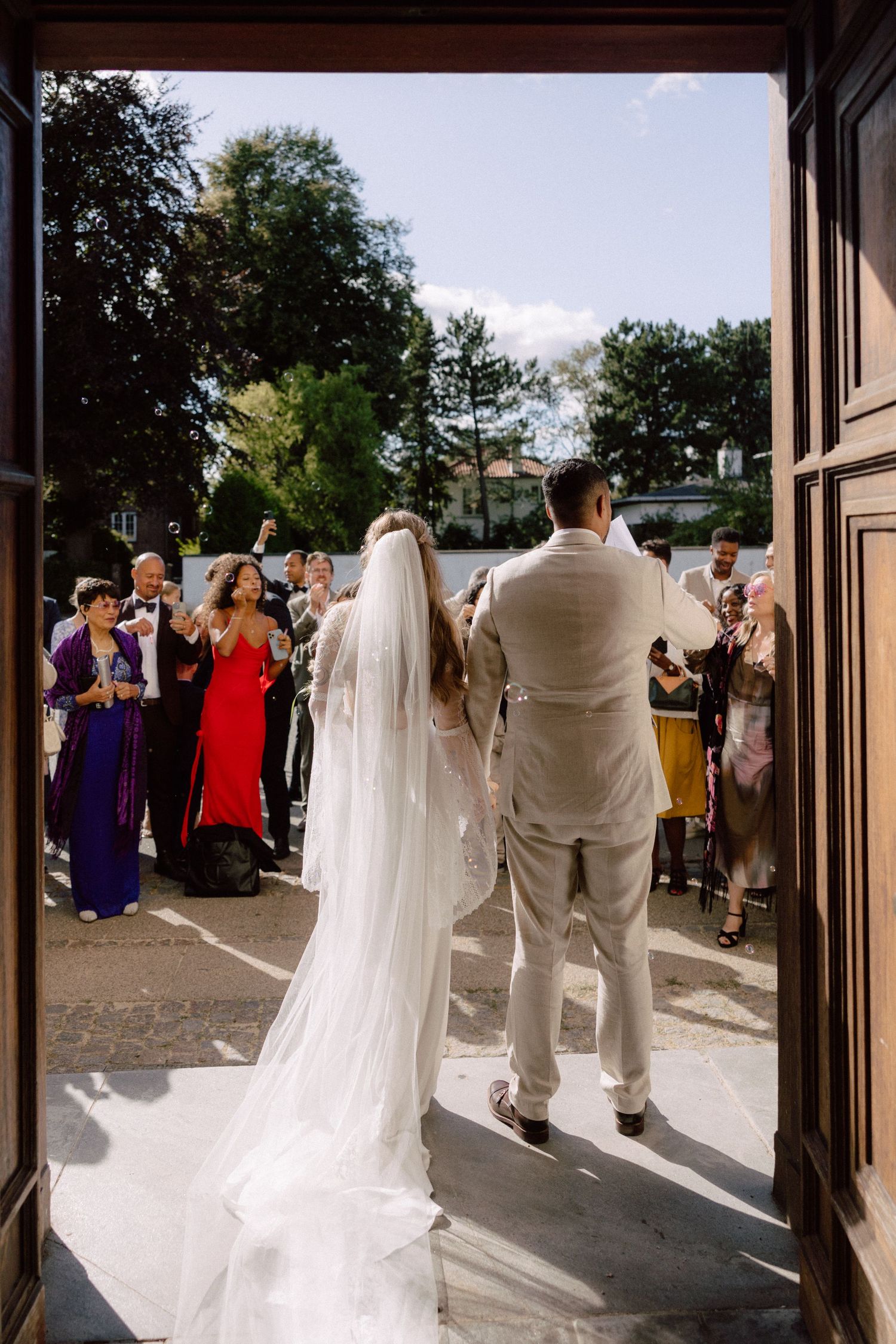 Wedding party and guests gather outside church doors in bright sunlight.