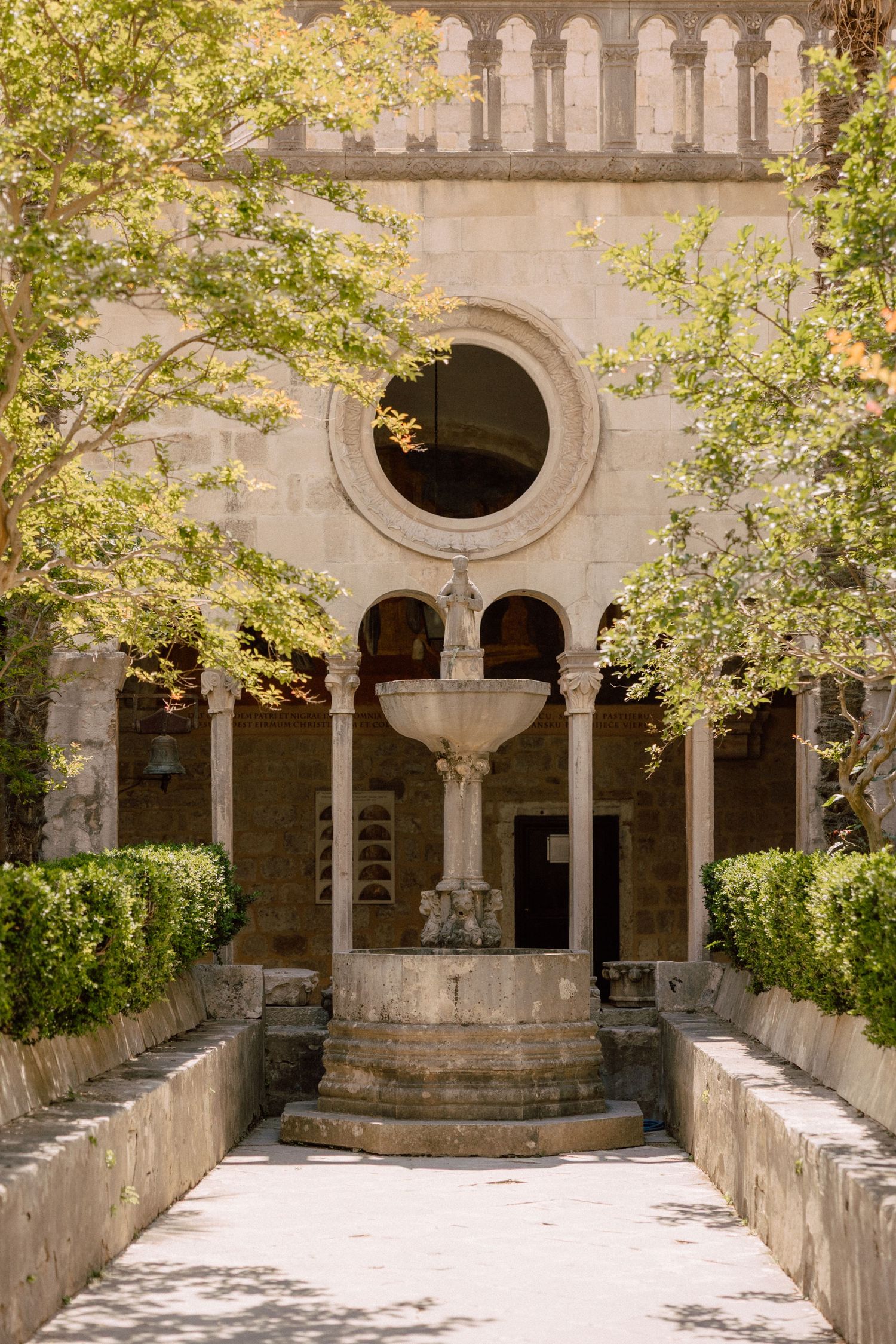 Historic monastery courtyard with stone arches and a central fountain surrounded by trees  in Dubrovnik, Croatia.