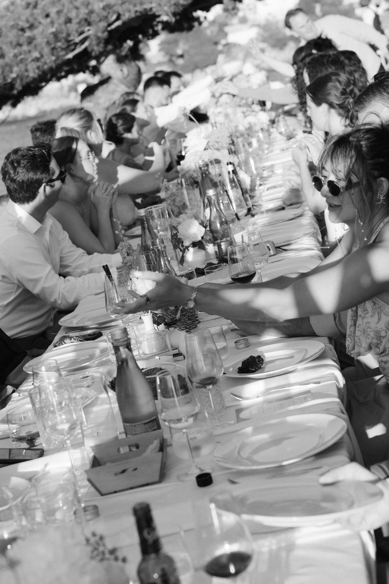 Black and white photo of guests dining at a long outdoor banquet table set with glasses, plates, and bottles.