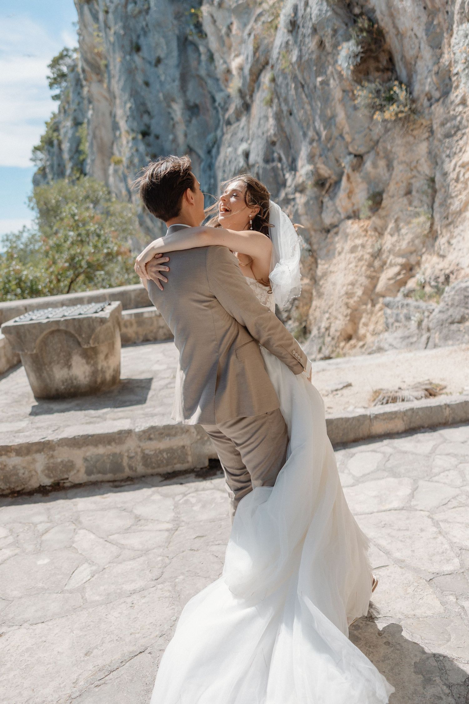 A joyful bride and groom embrace outdoors near rocky cliffs on their wedding day, with the bride in a flowing white gown.