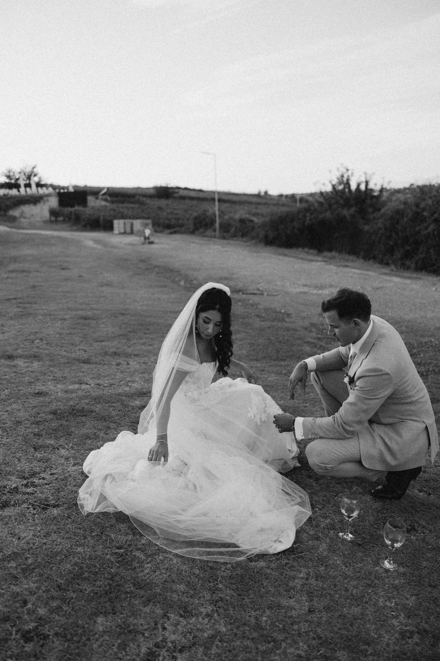 Wedding couple relaxes on grassy hillside with wine glasses at sunset.