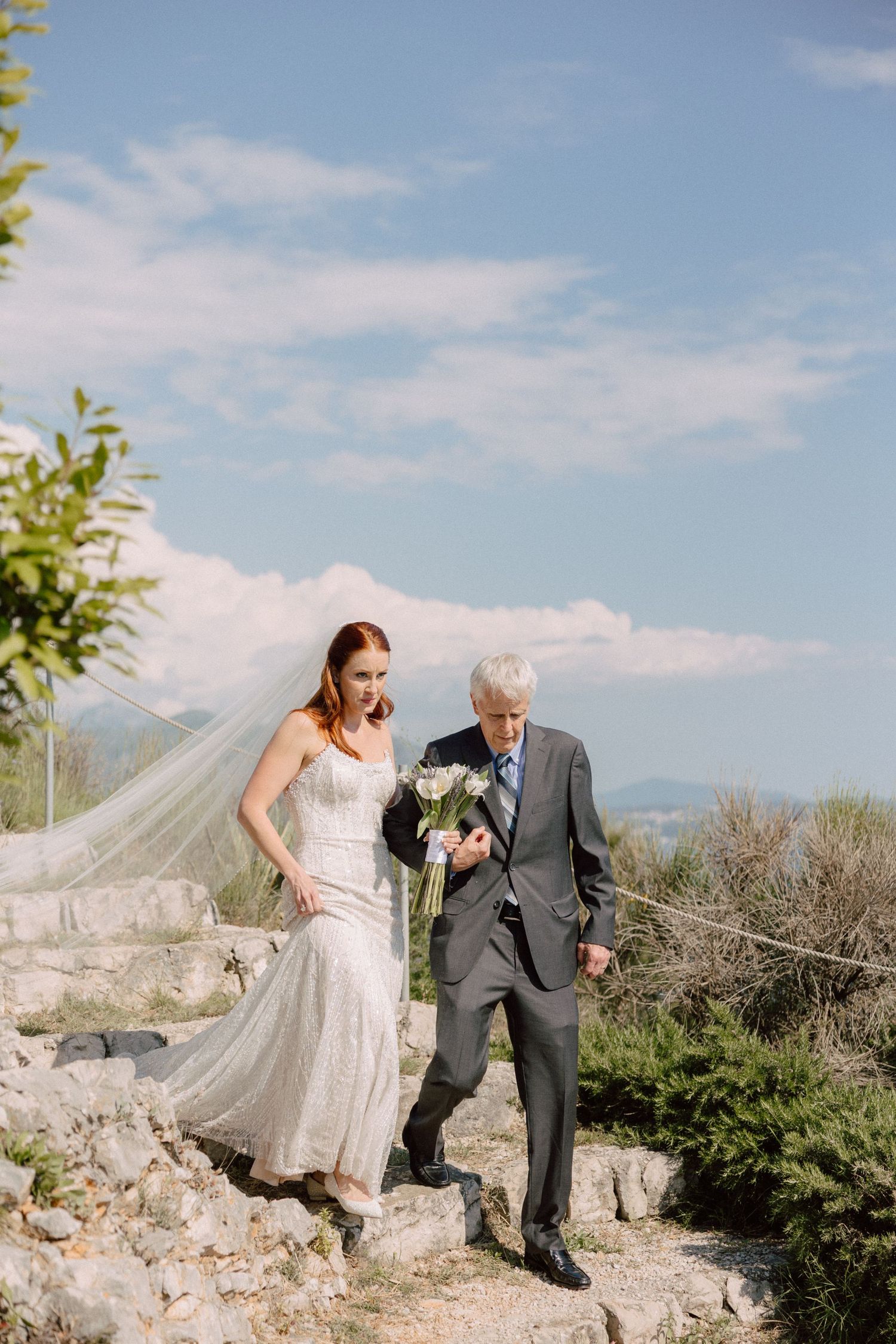 Bride and companion walking along a scenic mountain path on a sunny day with wispy clouds.