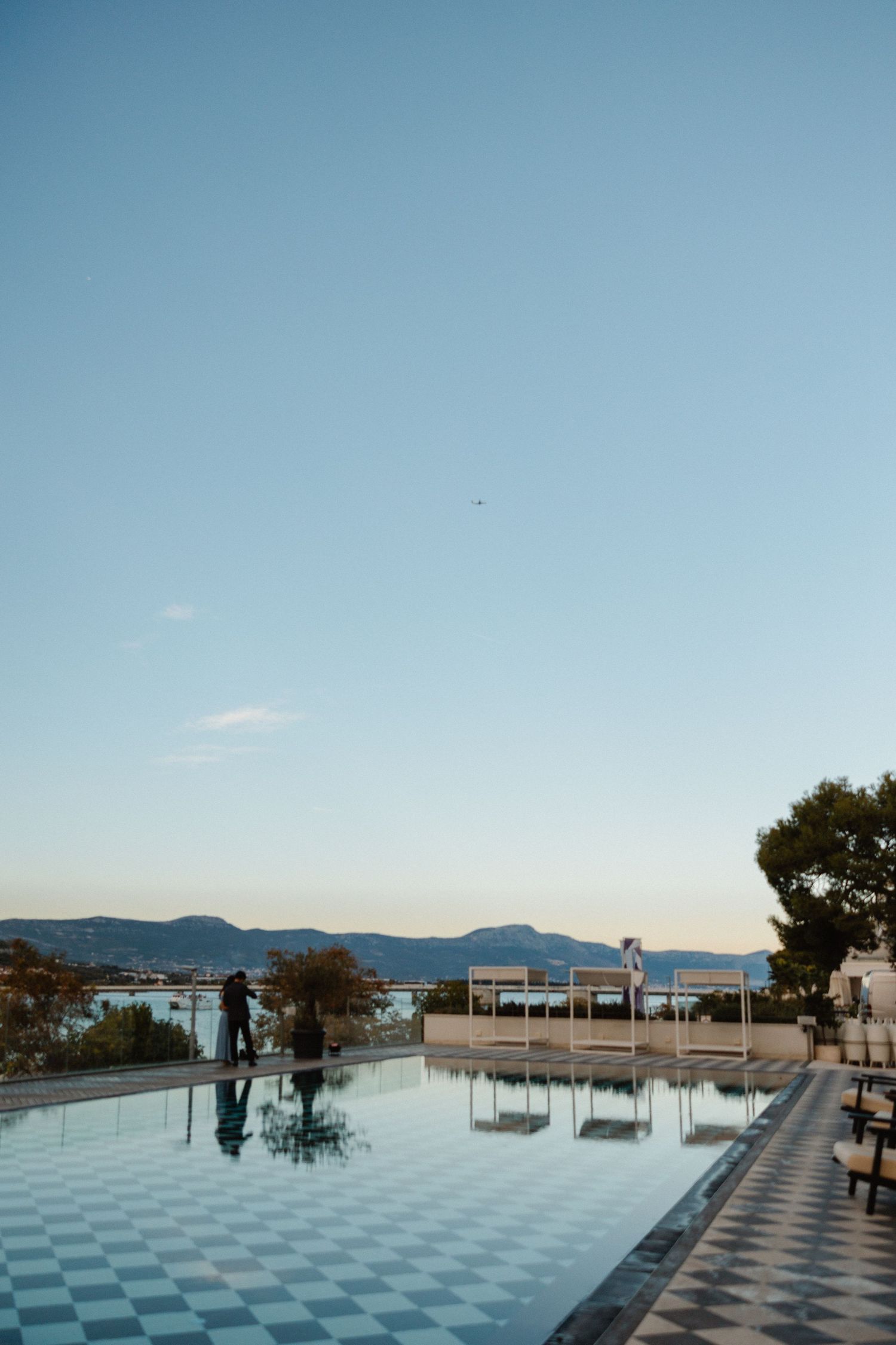 Couple stands by reflective pool at sunset with mountain and sea backdrop.