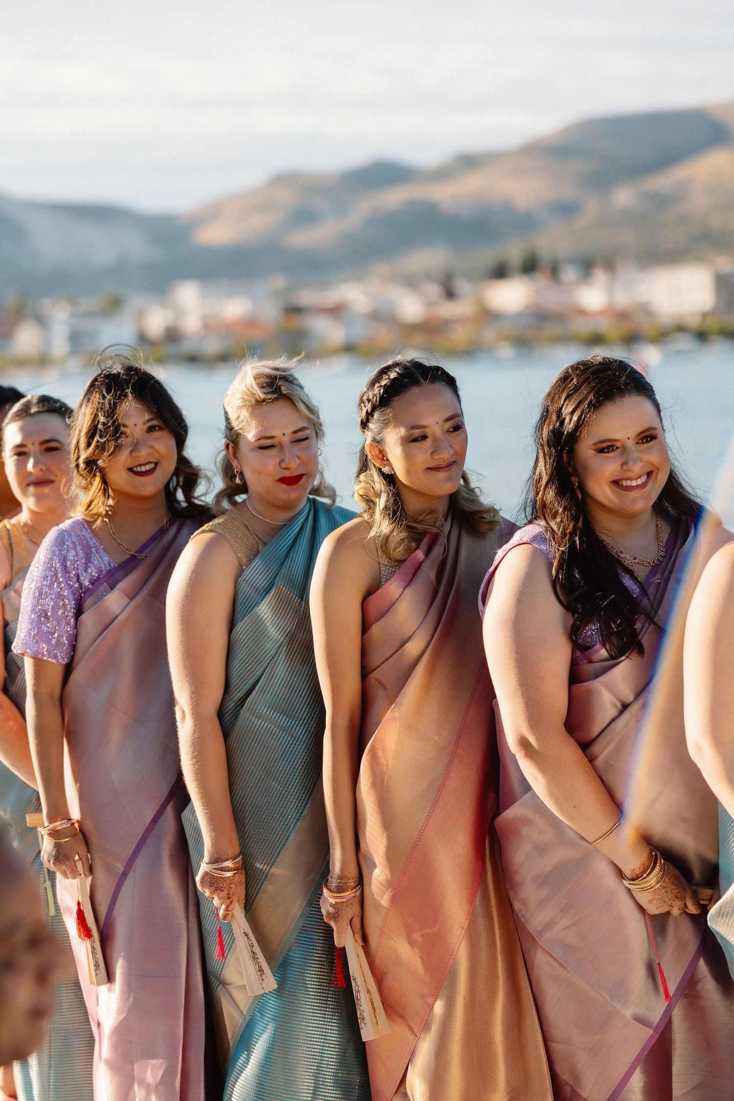 Bridesmaids in colorful silk sarees smile during outdoor waterfront wedding ceremony.
