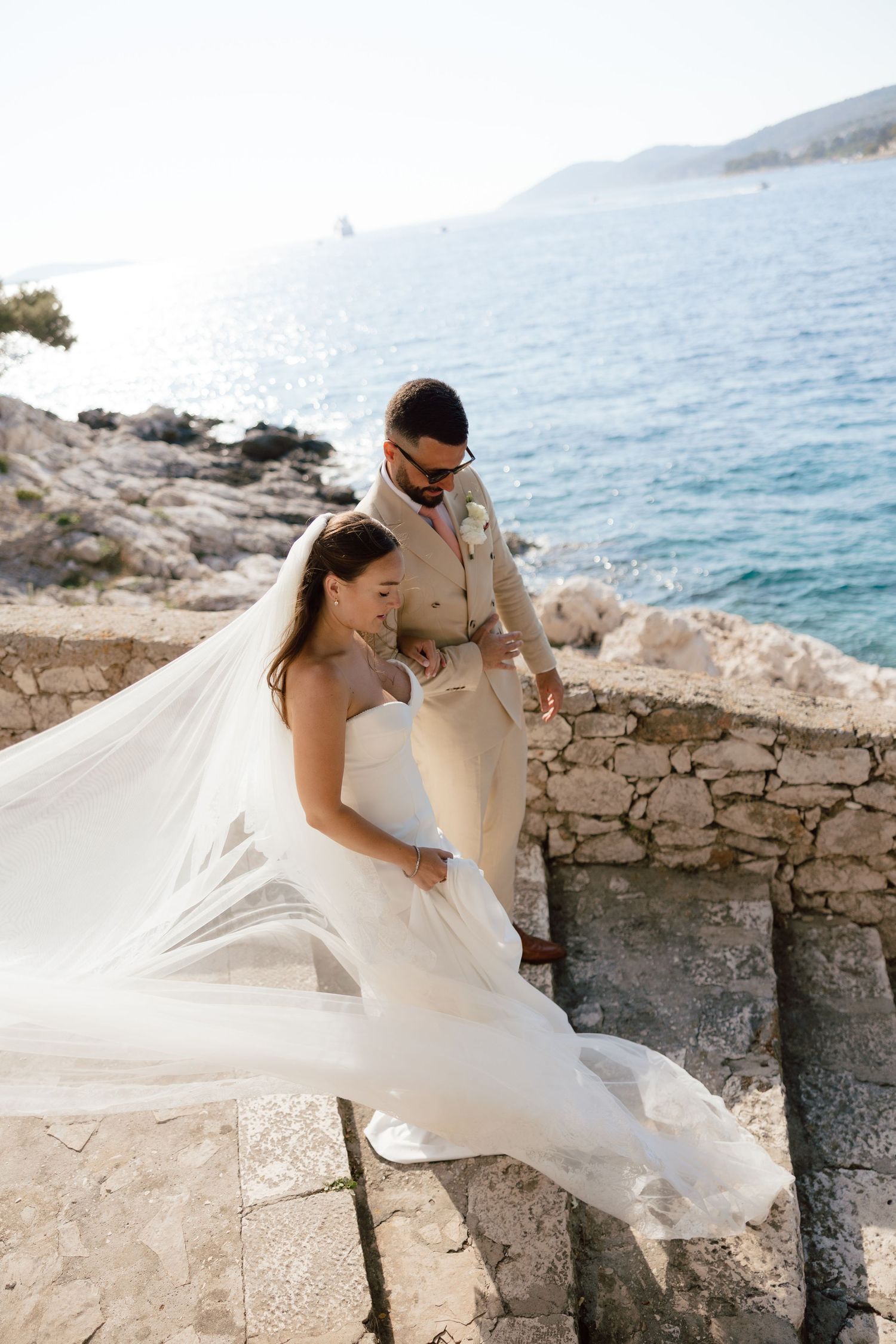 Bride and groom stand on stone wall with veil flowing by turquoise sea.