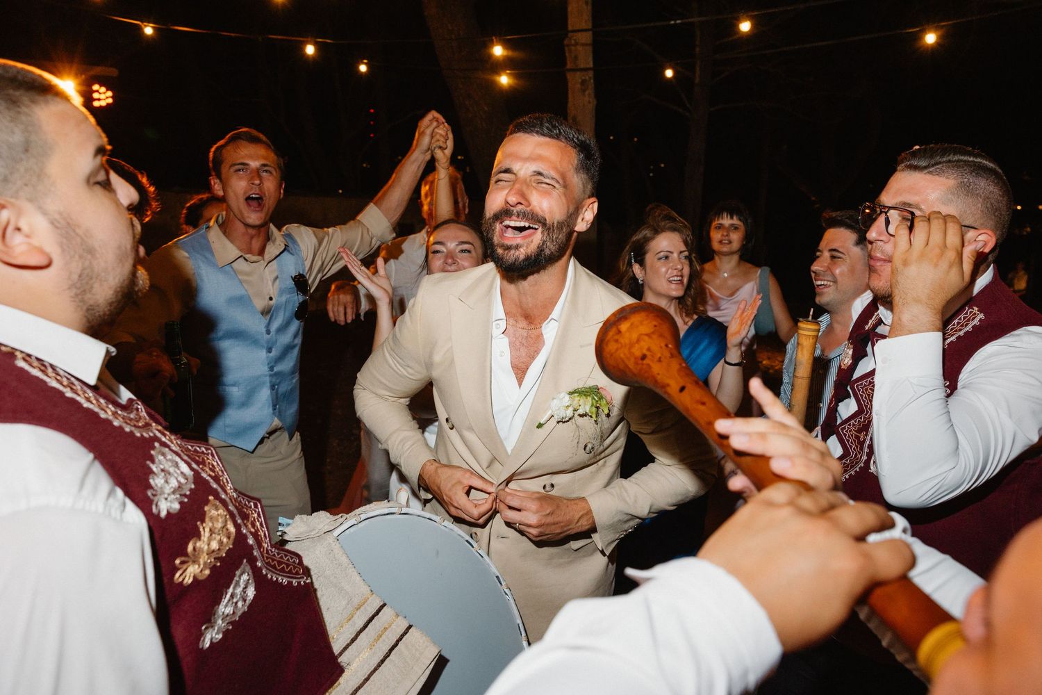 Groom in beige suit laughing and celebrating with traditional musicians at lively wedding reception.