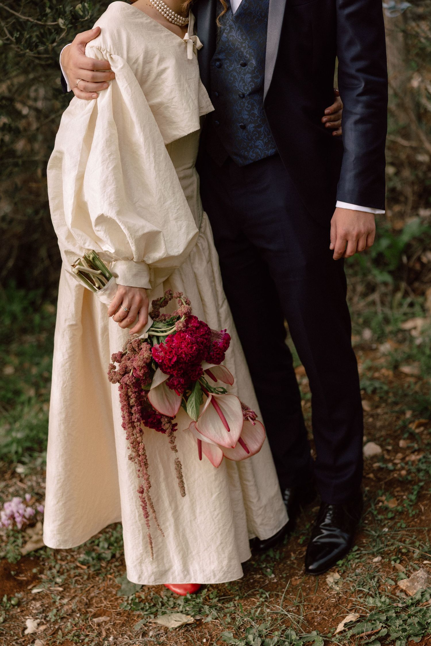 Wedding couple holding red bouquet against natural outdoor backdrop.