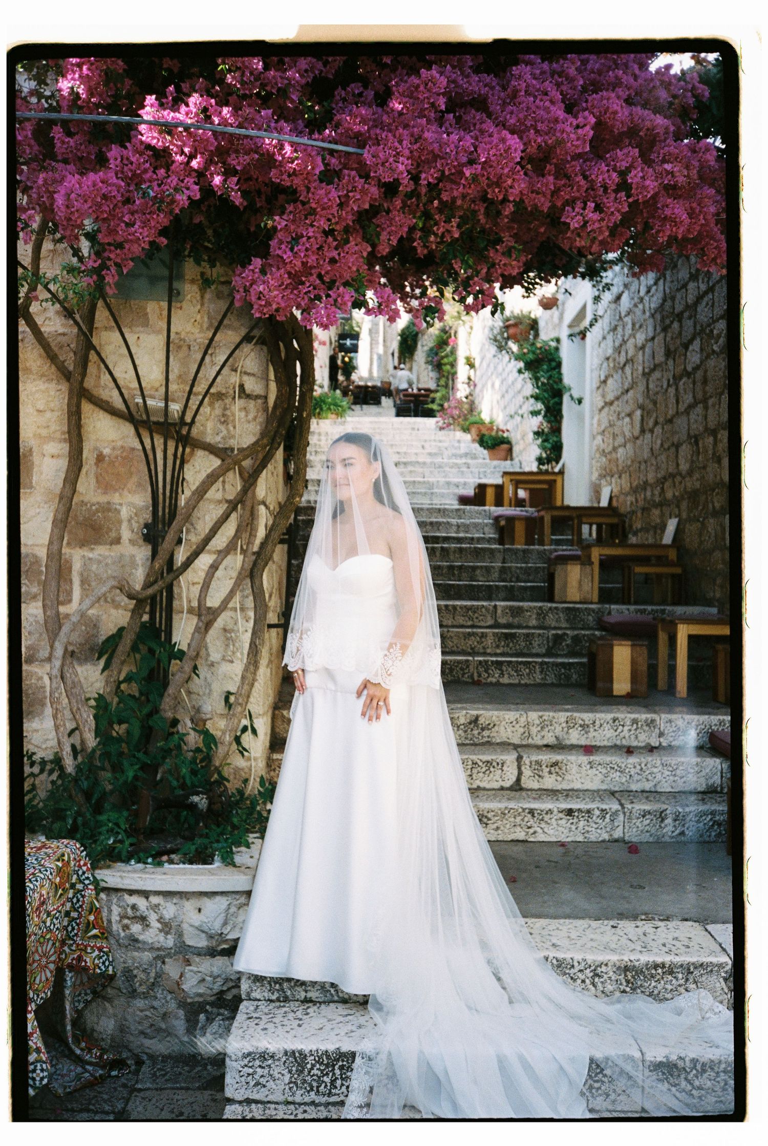 Bride in white gown and veil stands on stone steps beneath vibrant pink bougainvillea in a charming Mediterranean setting.