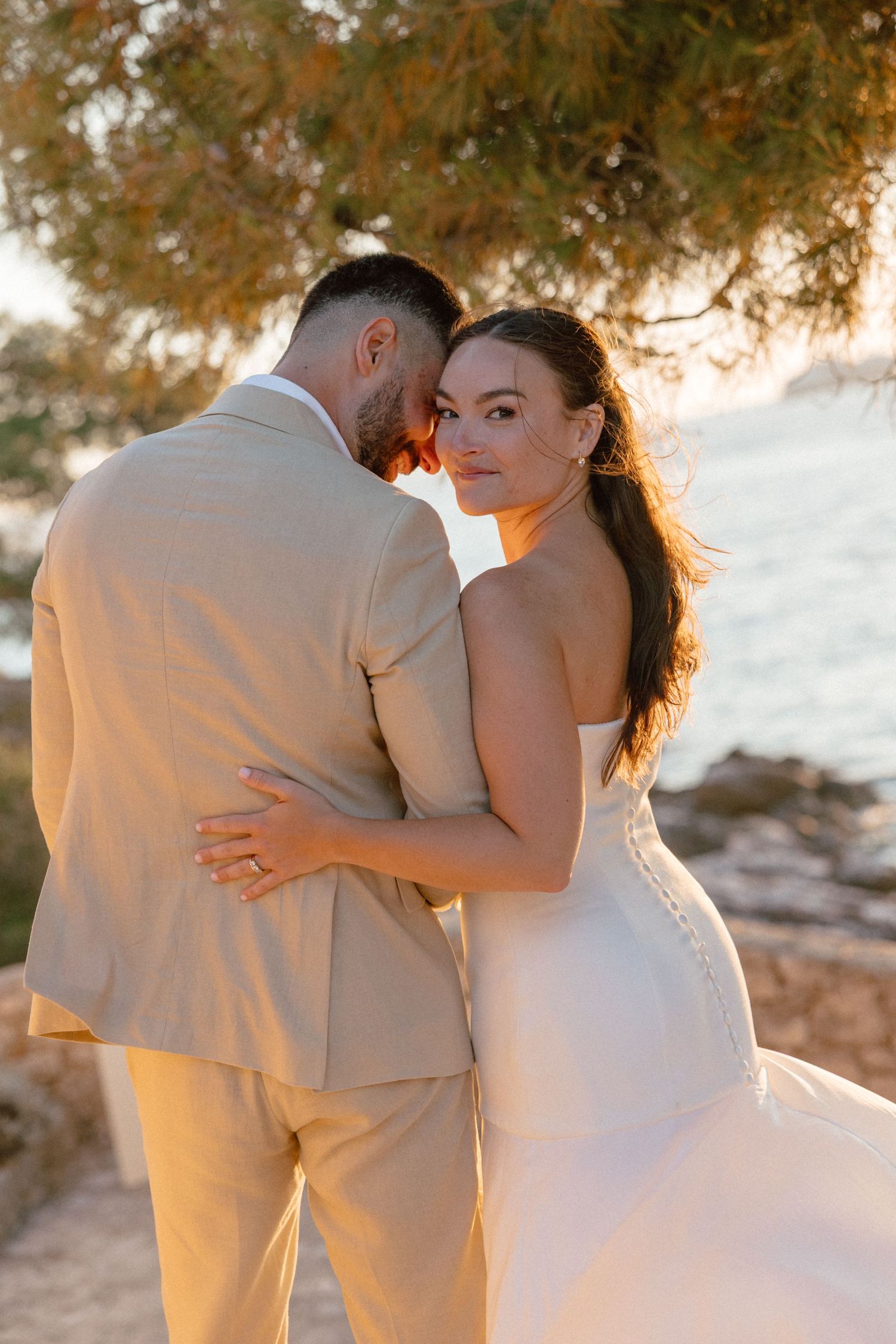 Bride in white strapless gown embraces groom in beige suit at golden hour by the sea.